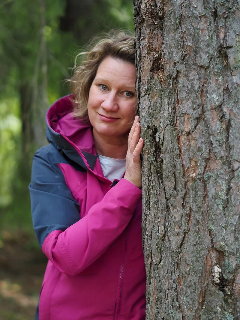 Toimintaterapeutti ja työnohjaaja Mia Pyykkö nojaa hymyillen puunrunkoon, pukeutuneena pinkki-siniseen takkiin.
