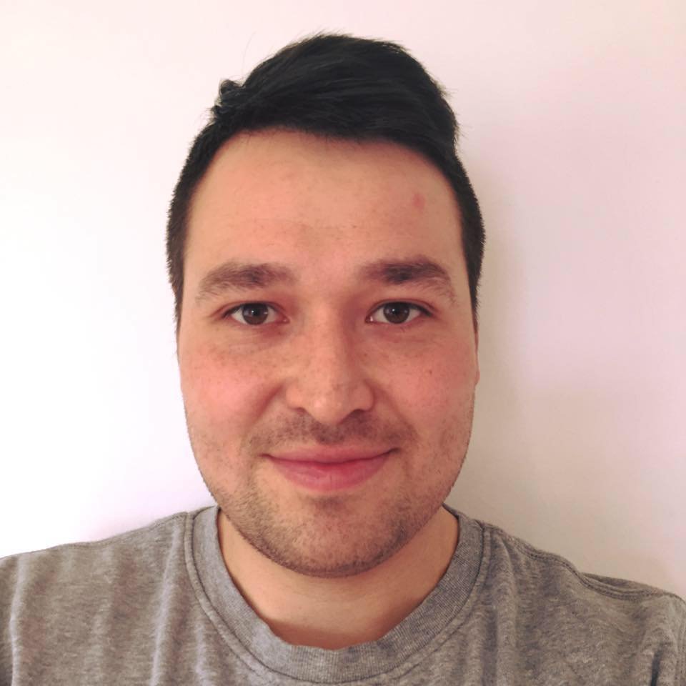 Portræt af en ung mand med sort hår, iført en grå T-shirt, der smiler mod kameraet foran en lyserød væg.