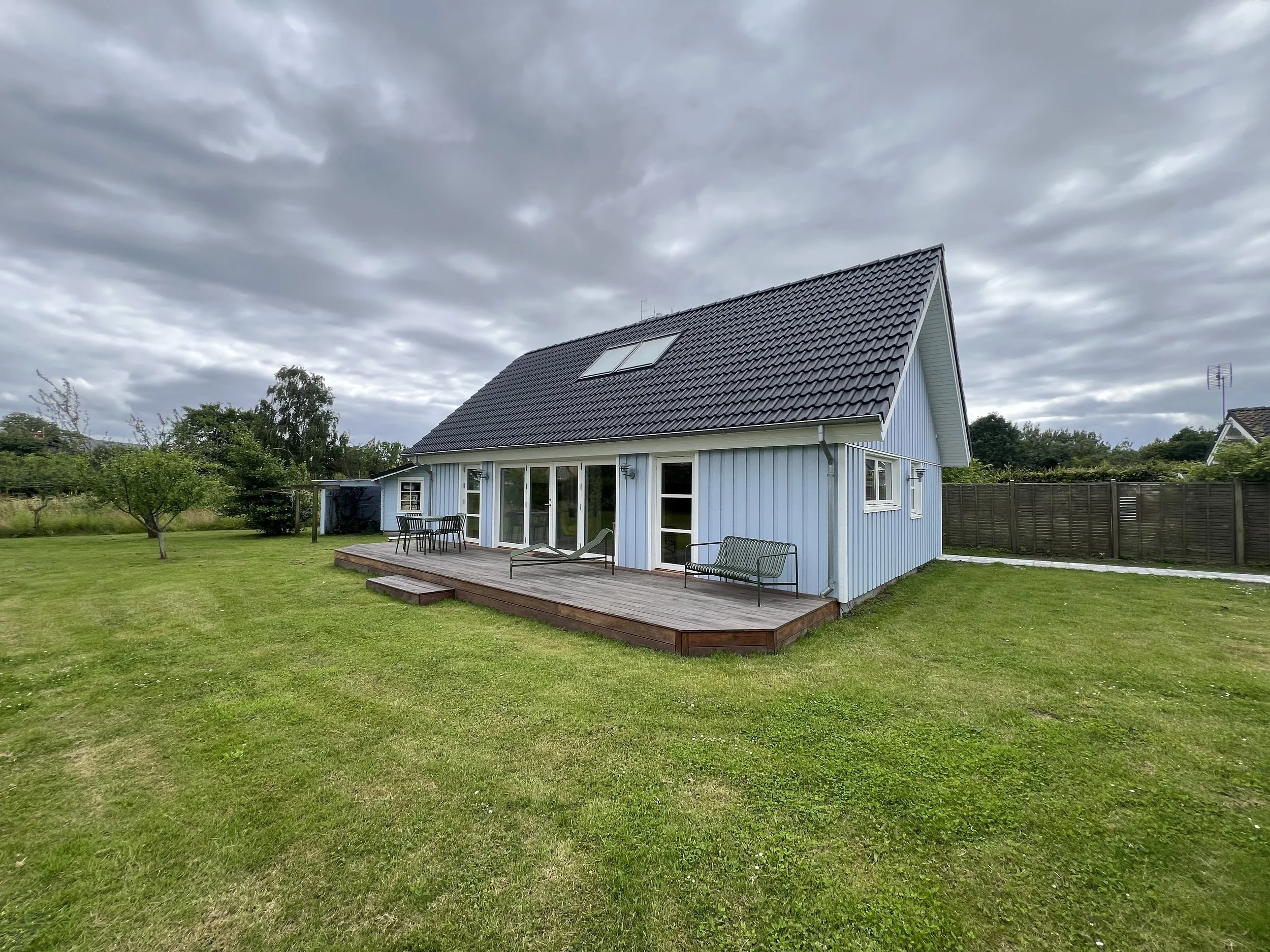Hus i lyseblåt med træterrasse og stor have, under overskyet himmel.