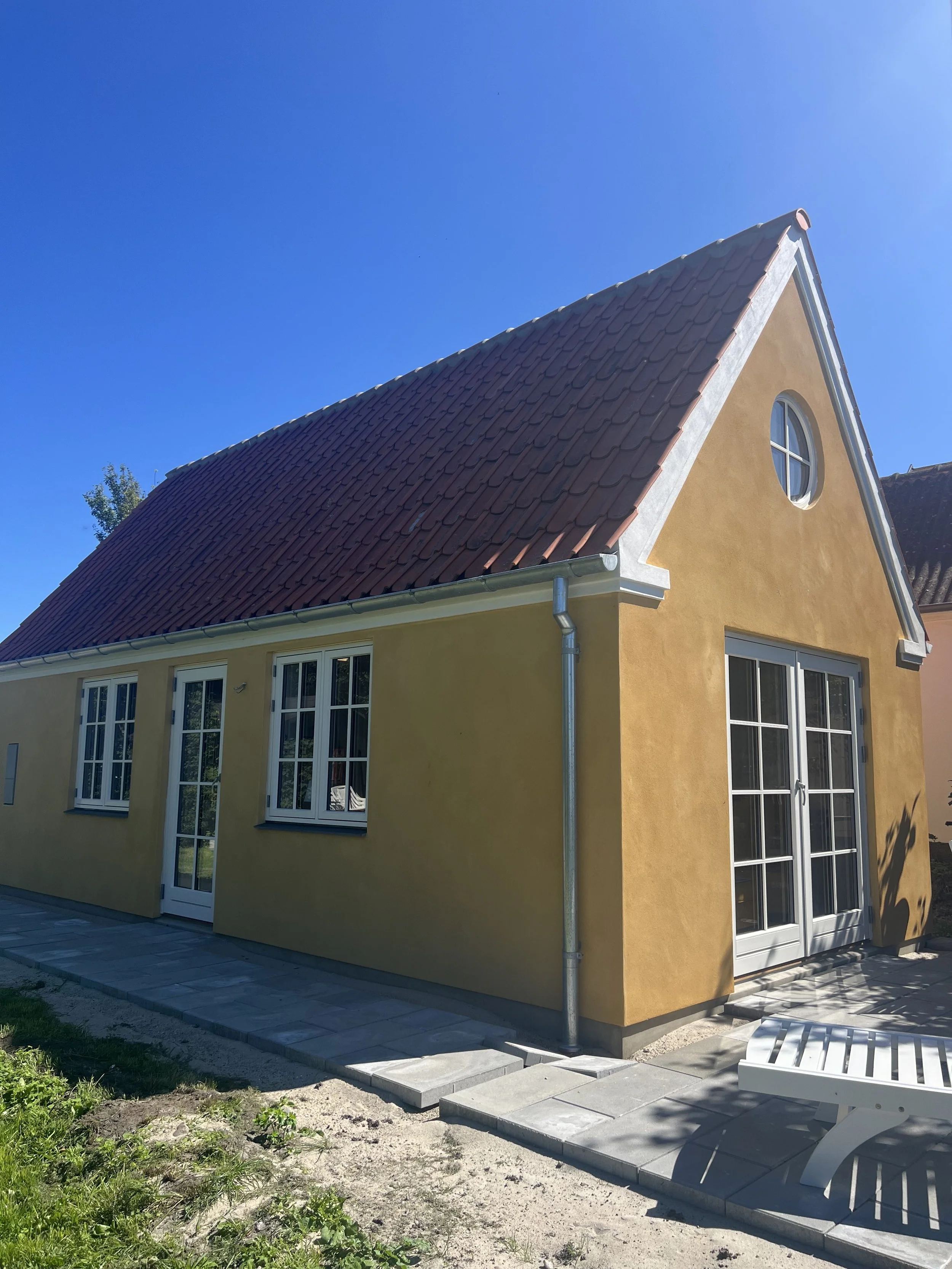 En gul, to-etagers hus med et rødt tegltag, hvide vinduer og en glasdør, med en grå flisebelagt terrasse foran.