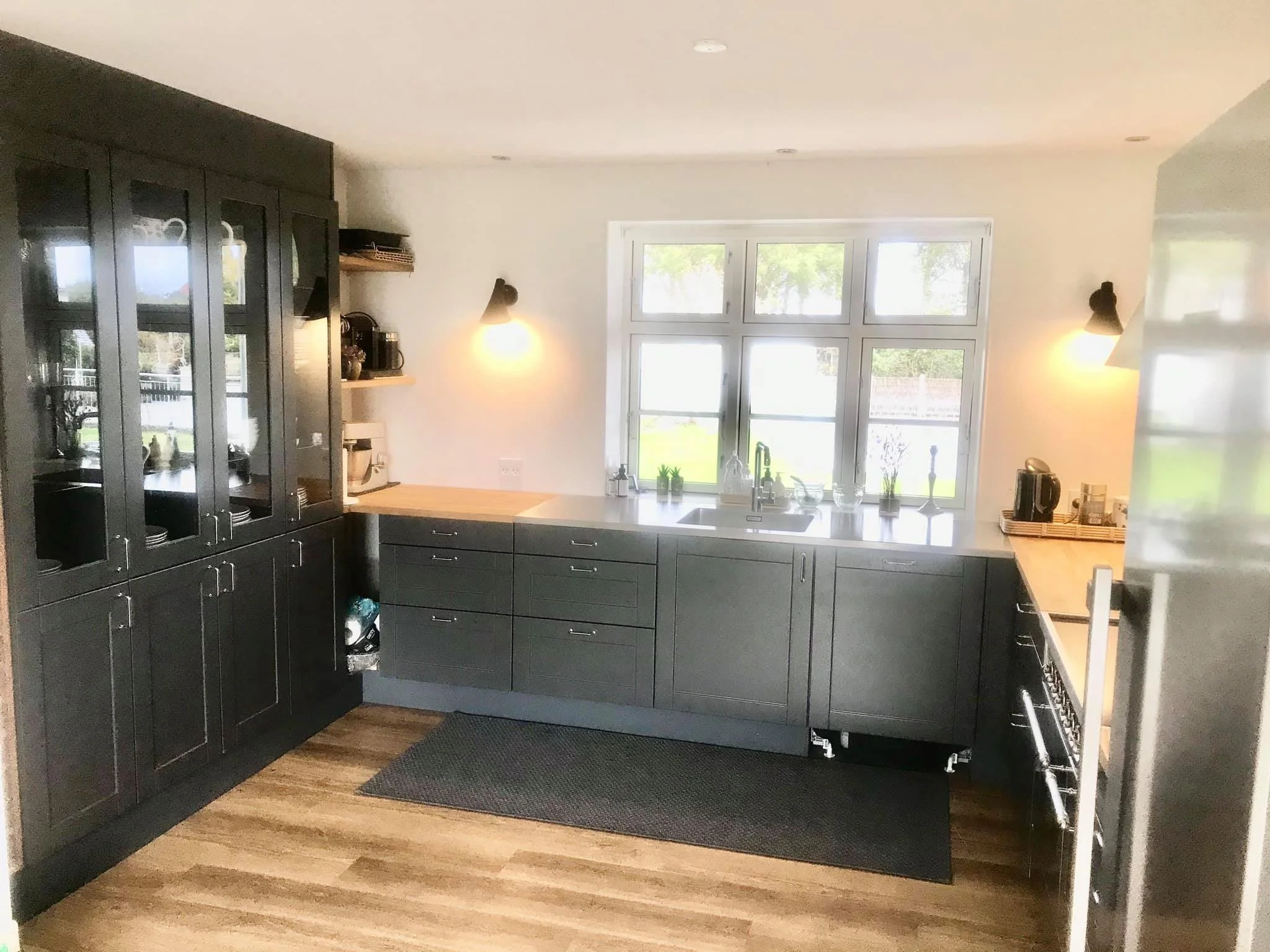 Køkken med sorte skabe, stor vindueskarm med planter og køkkenudstyr, træbordplade, vask under vindue, gasstof, og åbne hylder med kopper og apparater.