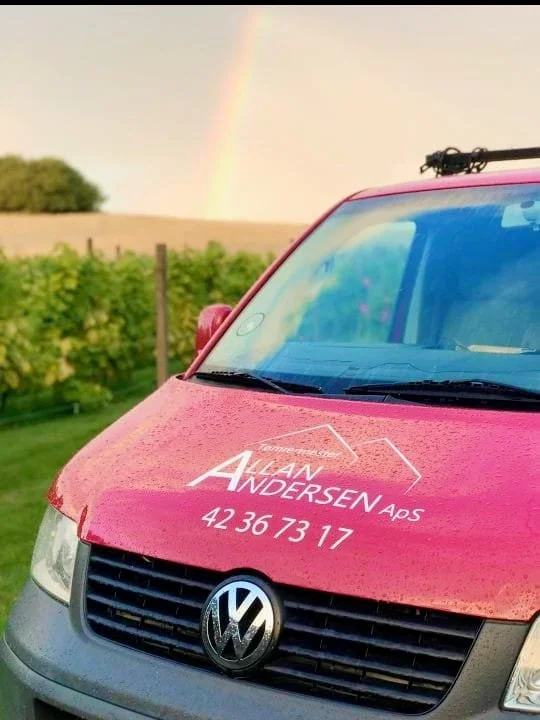 Pink Volkswagen varebil med logo og telefonnummer for ALAN ANDERSEN ApS, med regn på bilen og en regnbue i baggrunden