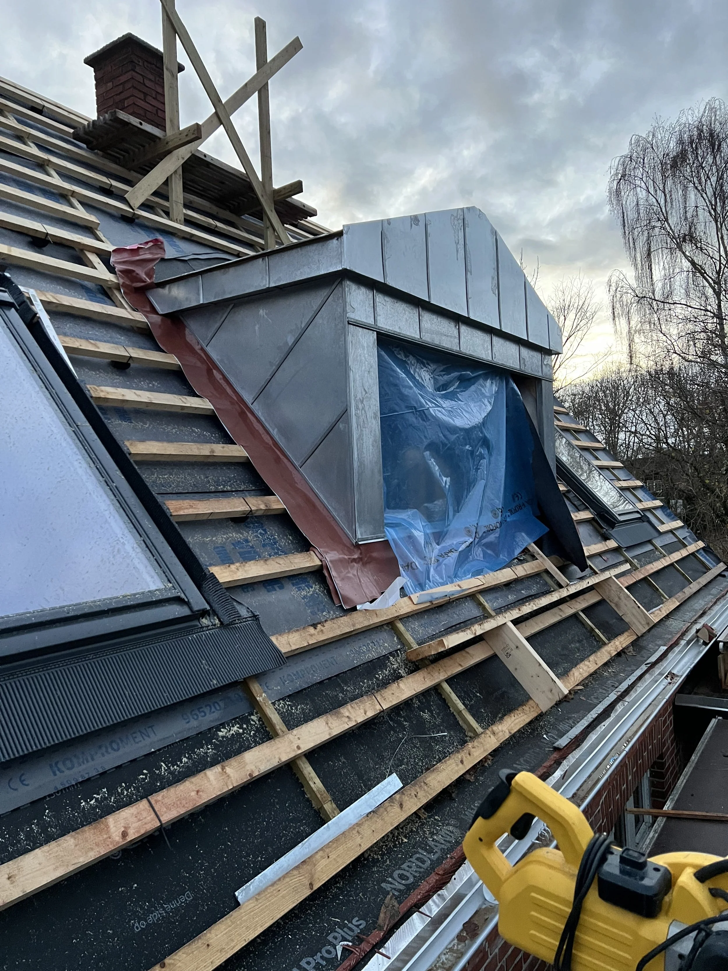 På taget af et hus er der under konstruktion med synlige tagsten og et nyt velux-udluftningsvindue, som er dækket af blå plastic for beskyttelse under tekniske installationer.