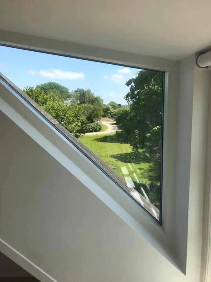 Udsigt gennem et skougt vindue med skrånende ramme, der viser grønne træer, en græsplæne og en sti udenfor.