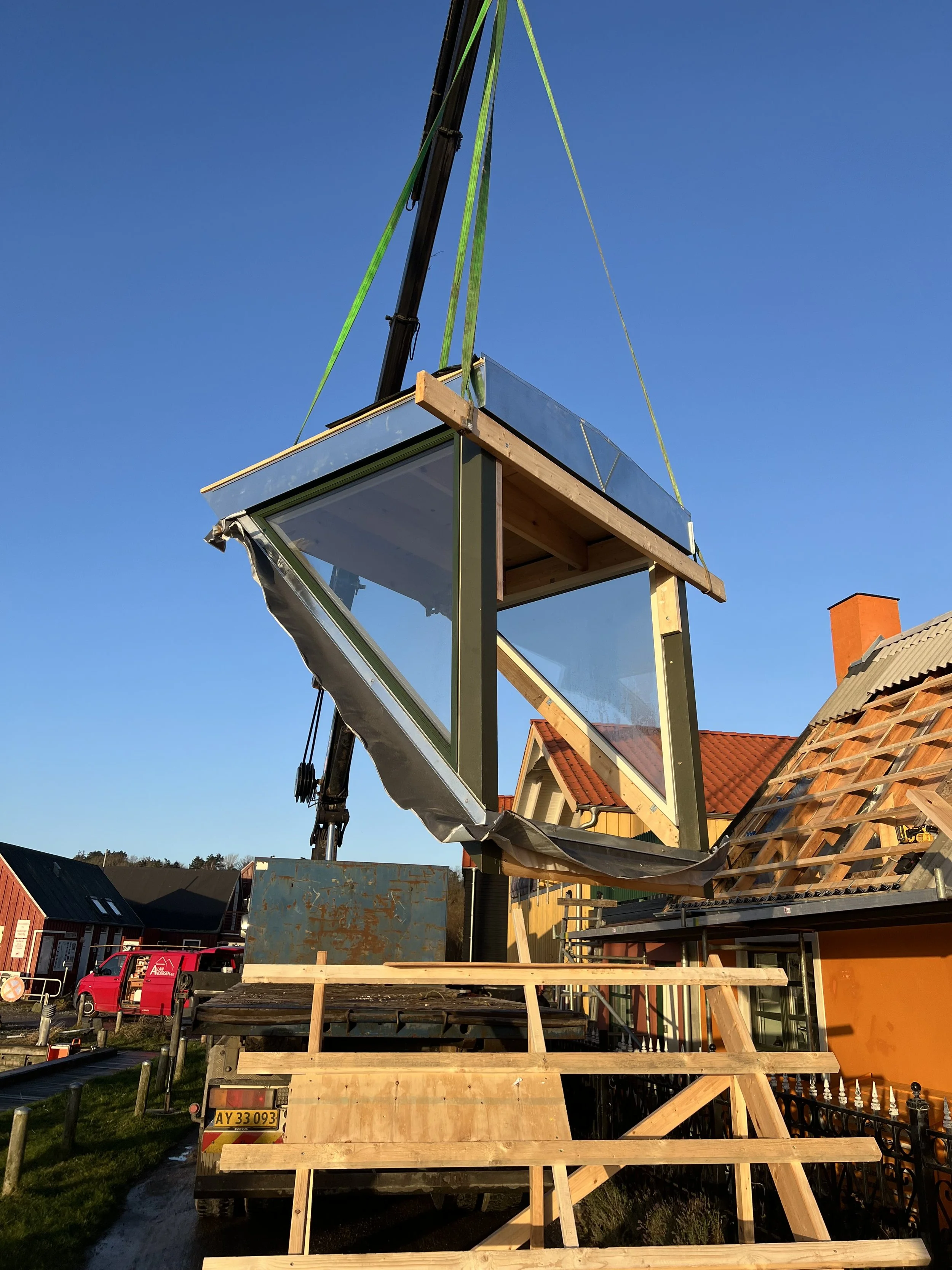 Et byggesæt med store glasvinduer hænges op af en kran foran et hus med rød tilstødende tage under en klar blå himmel.