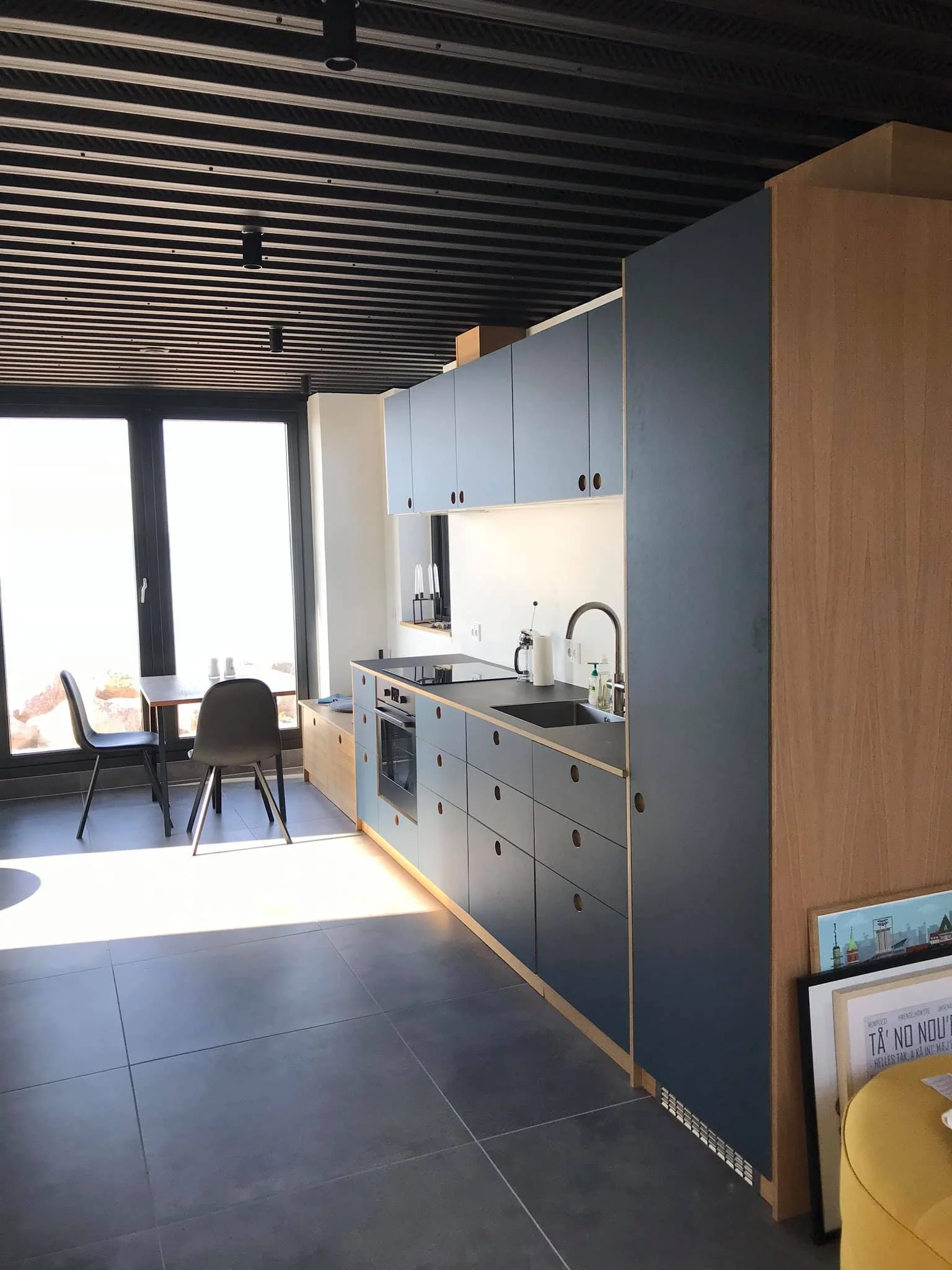 Moderne køkken med blå overhead- og skuffe-kabinetter, stor tilhørende vask, komfur, ovn, og gryder på bordet ud for vinduet. Der er et lille siddeområde med to stole ved et vindue.