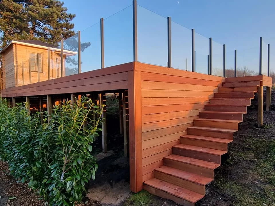 Terrasse med udendørs trapper, trægelænder og glasværn, tilstødende træhus i baggrunden, omgivet af grønne planter.