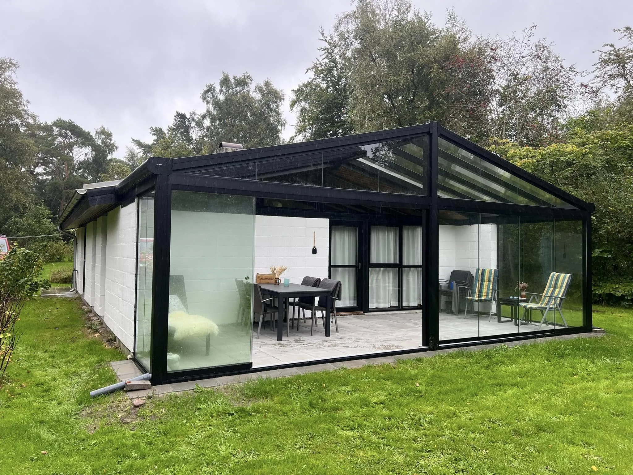 En glasindhegnet veranda med sort ramme og udendørs møbler i en grøn have.