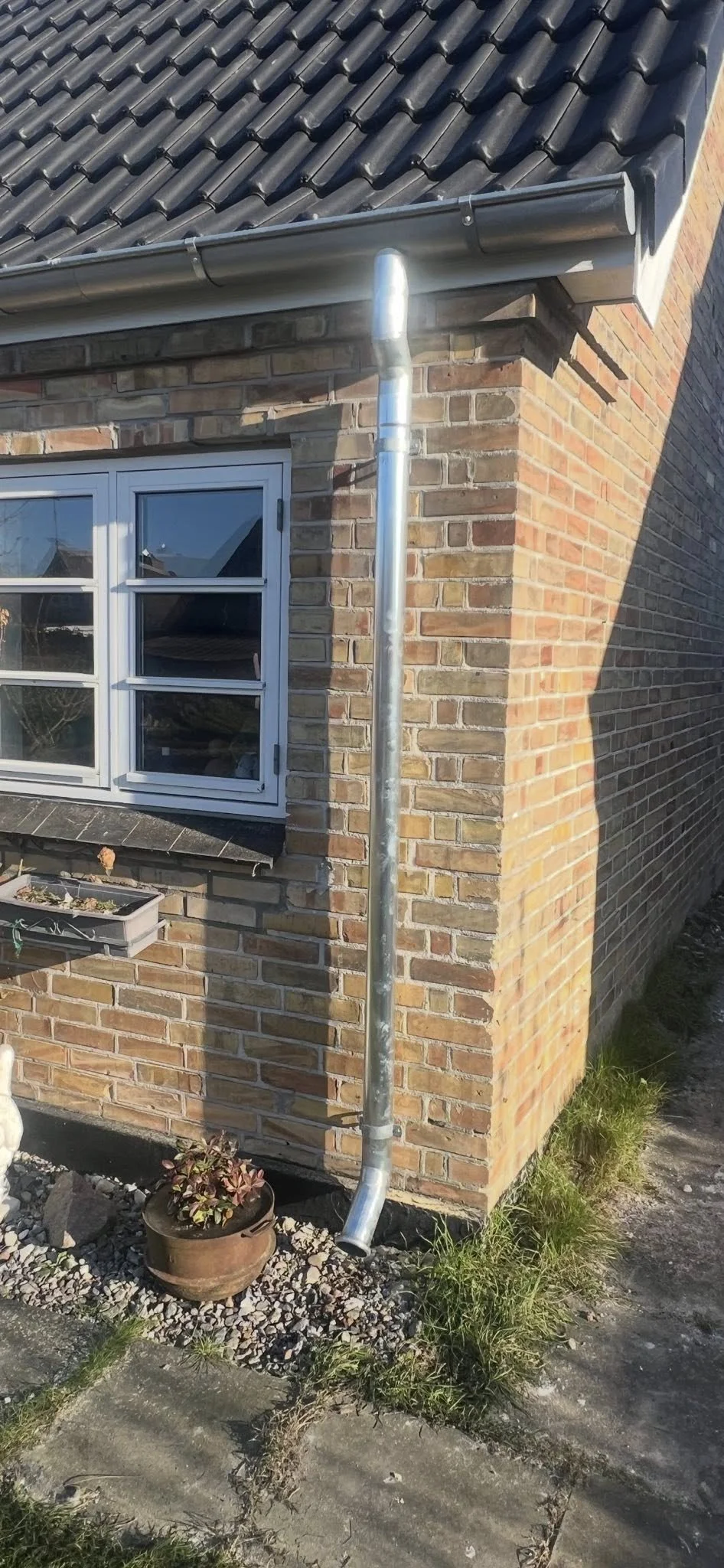 Et hjørne af en murstensbygning med et vindue, en tagrenderør, og en blomst i en potte på jorden.