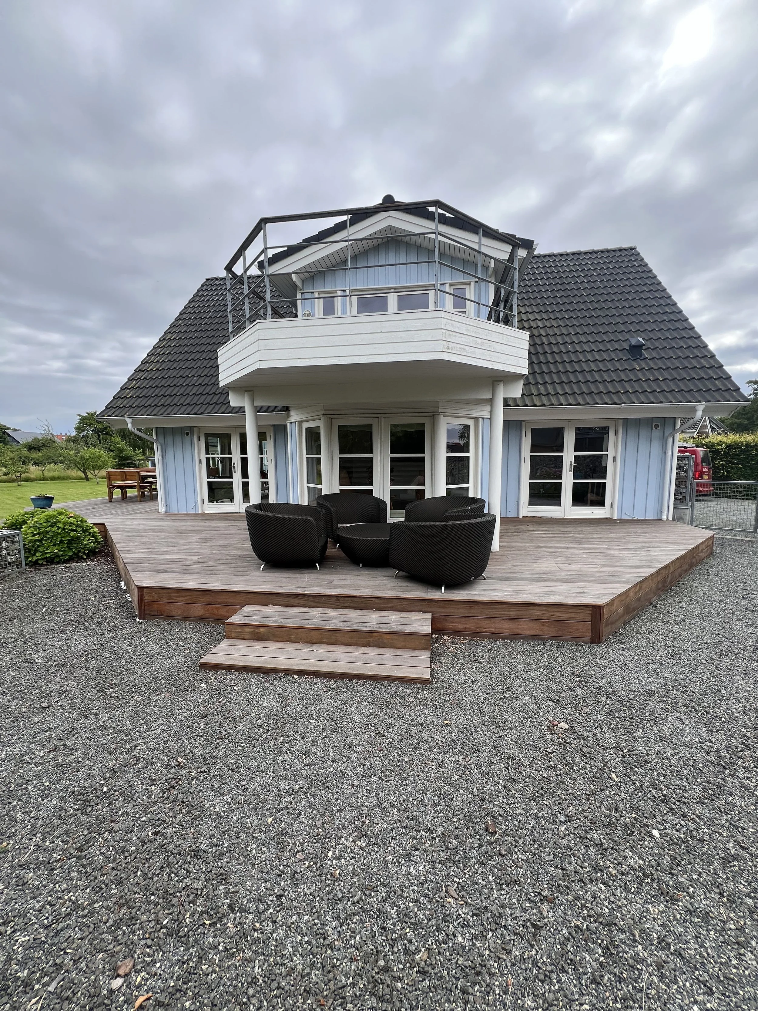 Et lyseblåt hus med stor træterrasse og udendørs siddeområde. Husets øverste balkon har metalræling, og der er græsplæne samt nogle planter i baggrunden. Overvejende overskyet vejr.