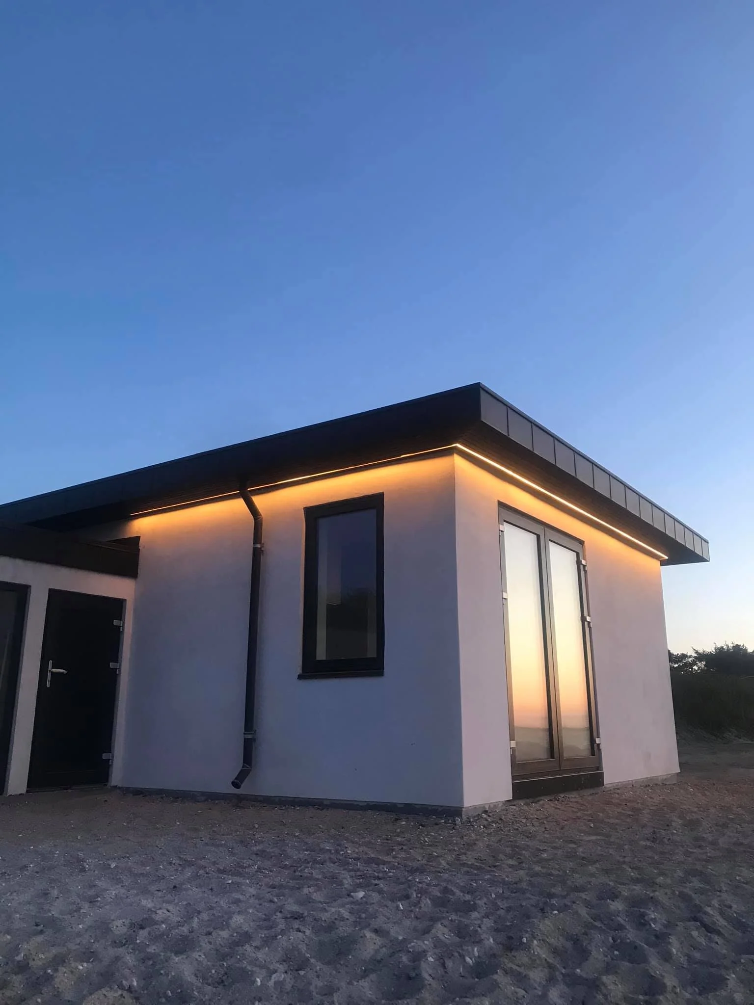 Moderne hytte med hvid facade, sorte vinduer og store glasdøre, set mod aftenhimlen med reflektion af solnedgang i glasset.