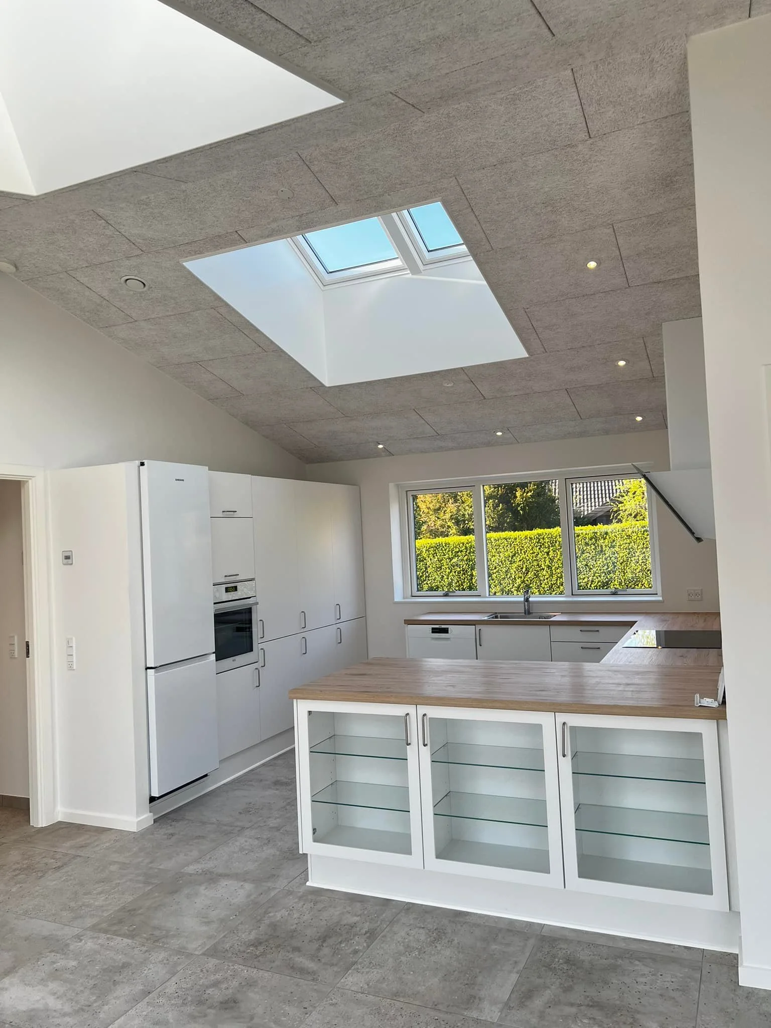 Et moderne køkken med hvid skabsindretning, træbord og store vinduer samt et ovenlys i loftet.