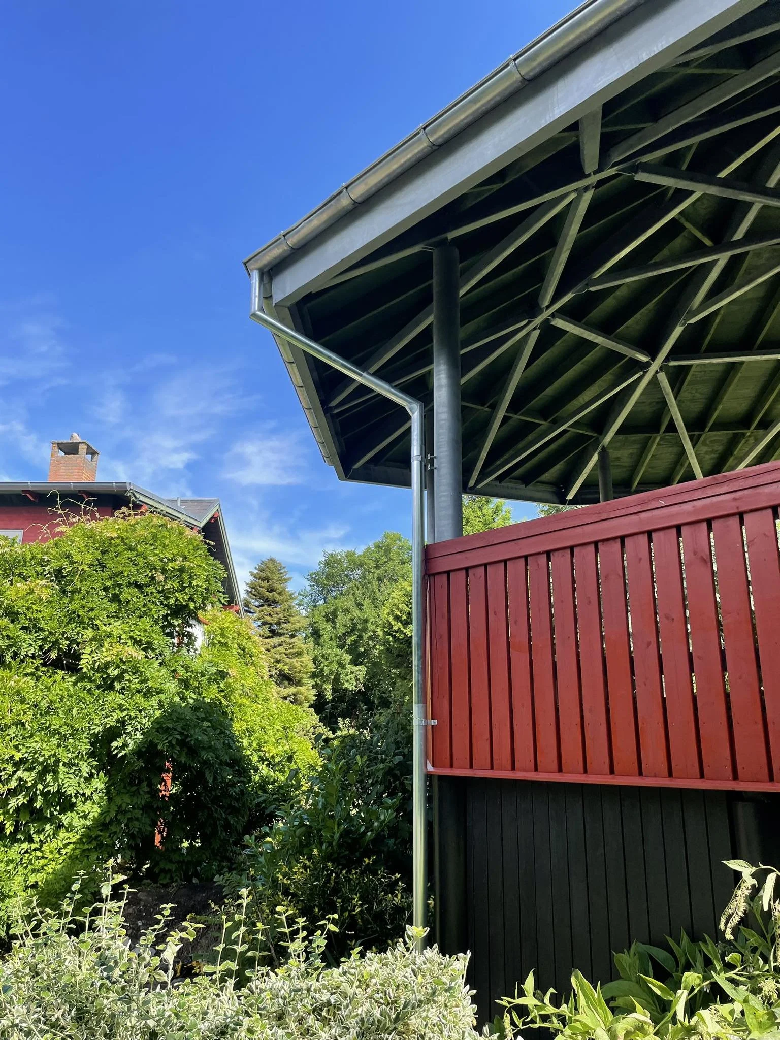 Et hjørne af en bygning med et underliggende tag, en rød træskærm, grønne buske og træer, blå himmel og en skorsten på en anden bygning i baggrunden.