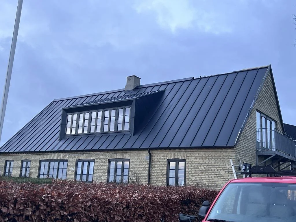 Et hus med gul/brun mursten, store vinduer og en sort, metal tag. Der er en balkon på øverste etage.