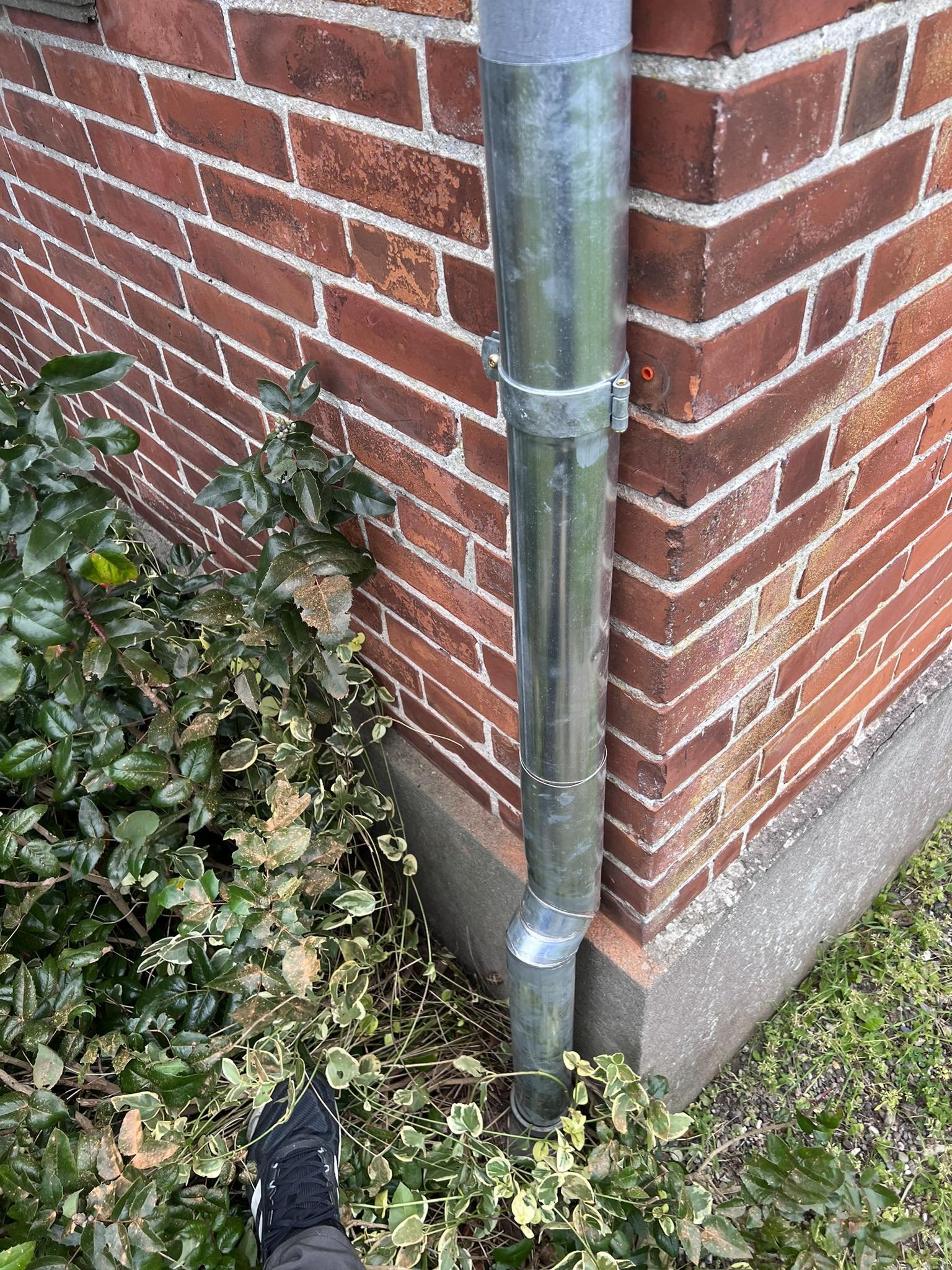 En hjørne af en bygning med en rød murstensmur og en metalskorsten, omgivet af grønne planter.