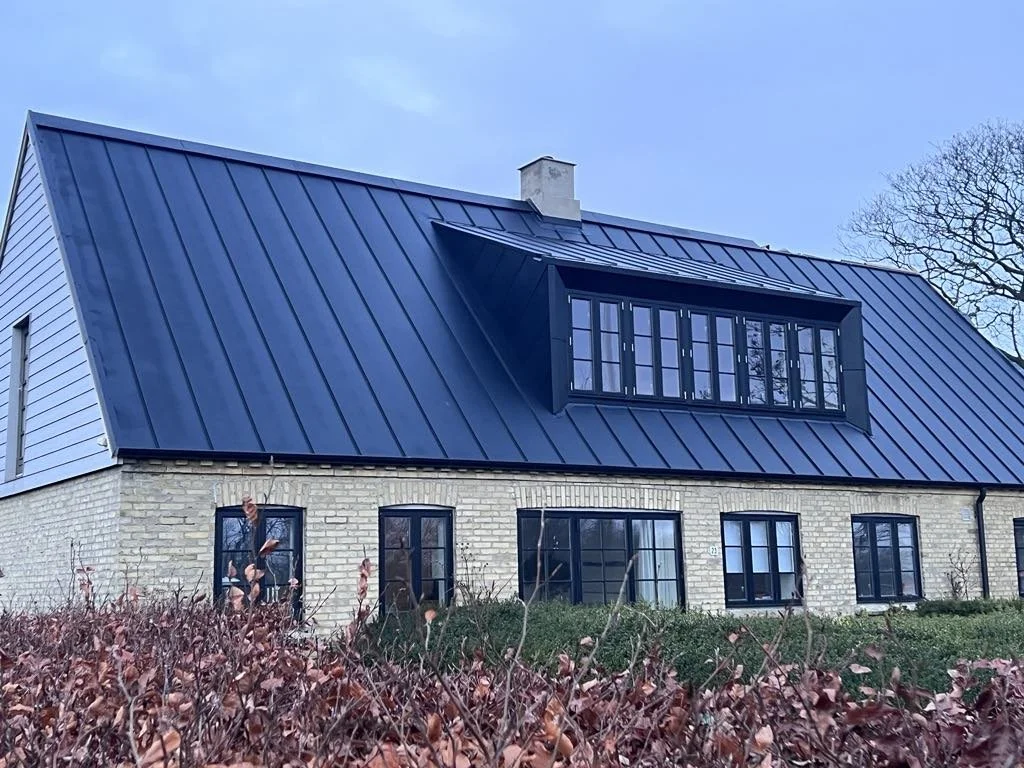 Et hus med en blåmetaltag, to vinduer i stueetagen og et større opbevaringsvindue på loftet. Der er buske foran huset og en klar himmel.