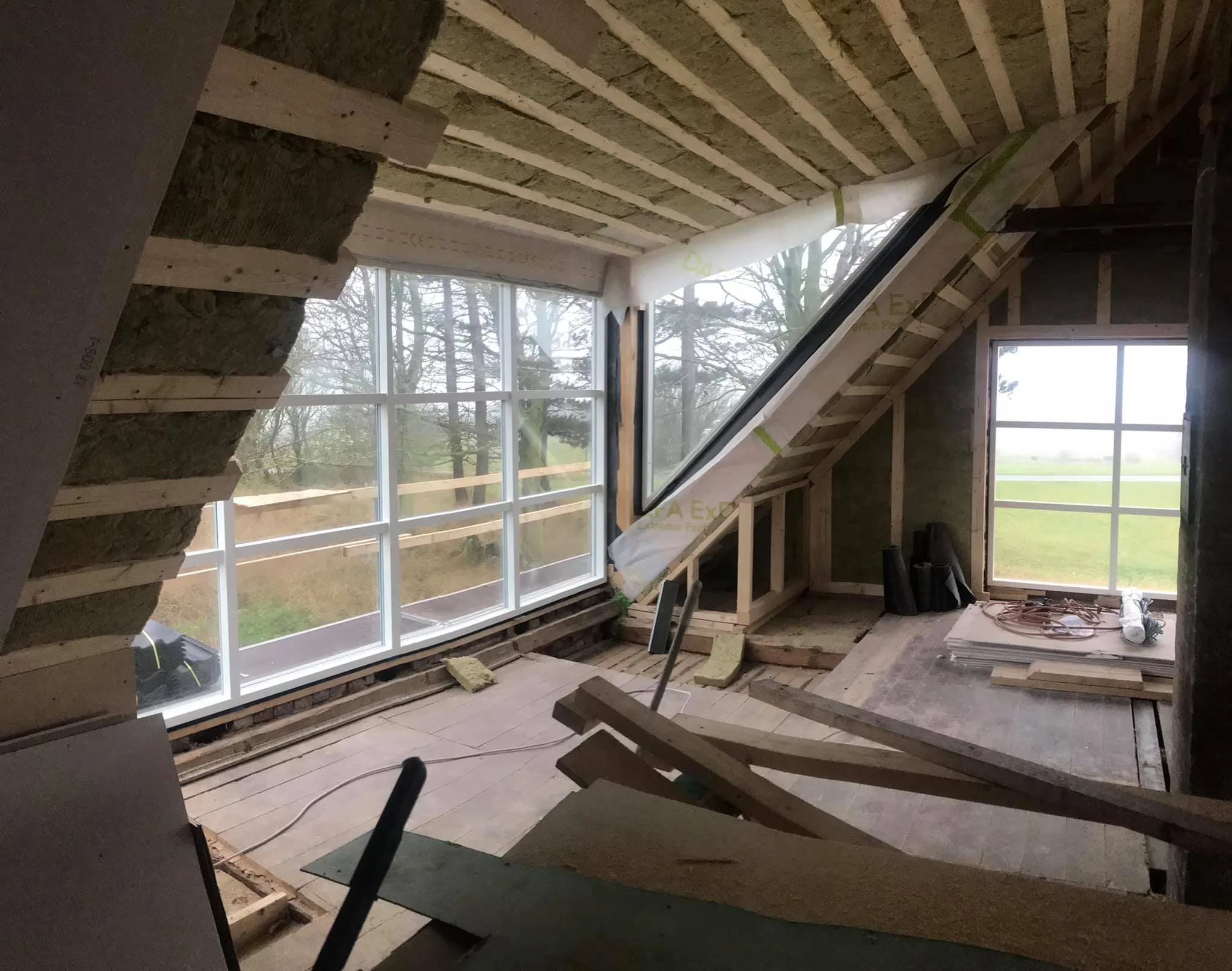 Under konstruktion: et rum med store vinduer, der vender ud mod en grøn have, med synlige træbjælker, isolering og byggematerialer.