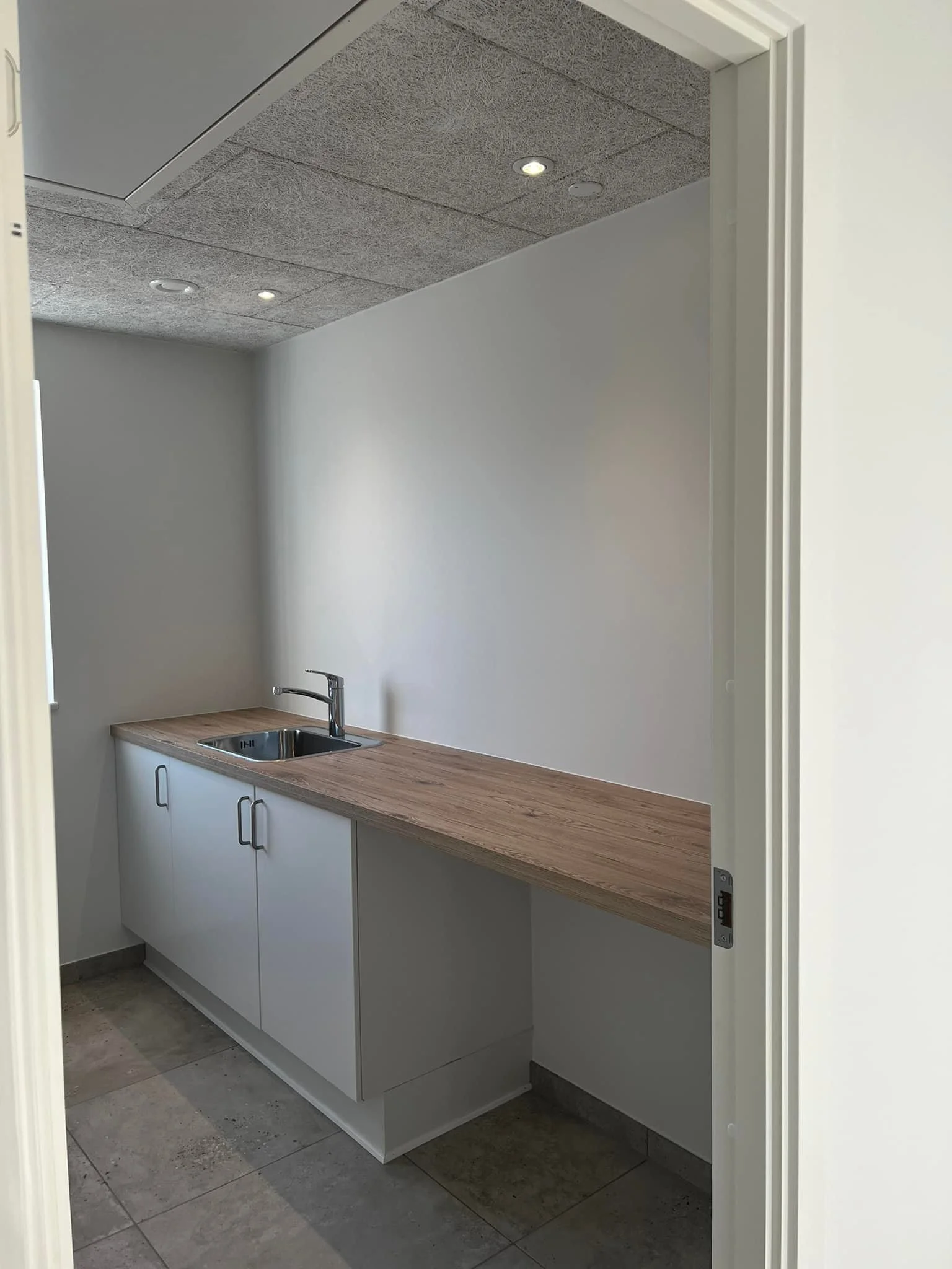 Et lille køkken med hvidt skab under vasken, en træbordplade og en enkelt vandhane, i et rum med hvide vægge og en loftsflade med indbyggede lys.