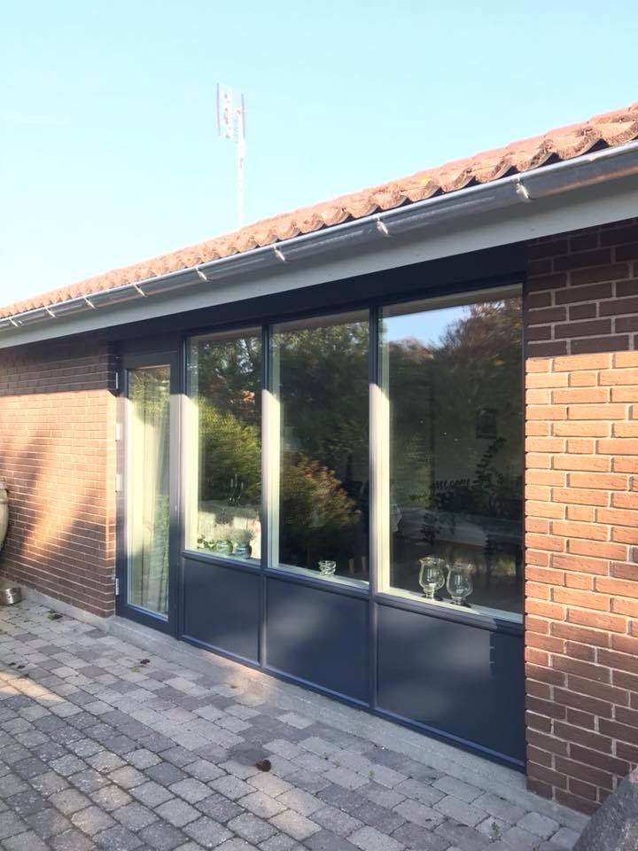 Udendørs udsigt af en del af en bolig med store vinduespartier, en mur af røde mursten, og et tag med tagsten, under en klar blå himmel.