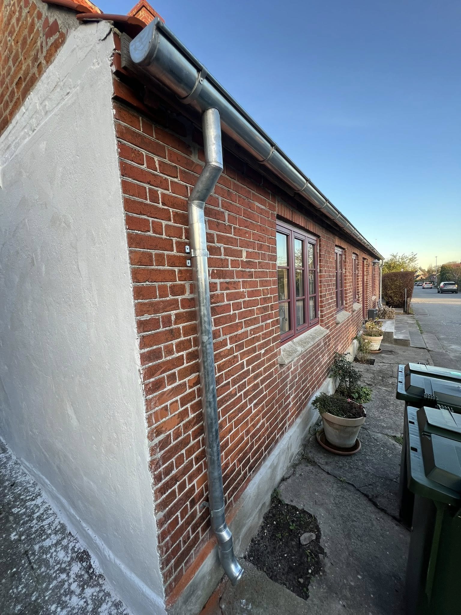 Et udendørs billede af et rødt murstenshus med to vinduer og metalrør, der fører regnvand væk fra tagrenderesystemet. En beholder med planter står foran huset, og resten af gaden kan ses i baggrunden.