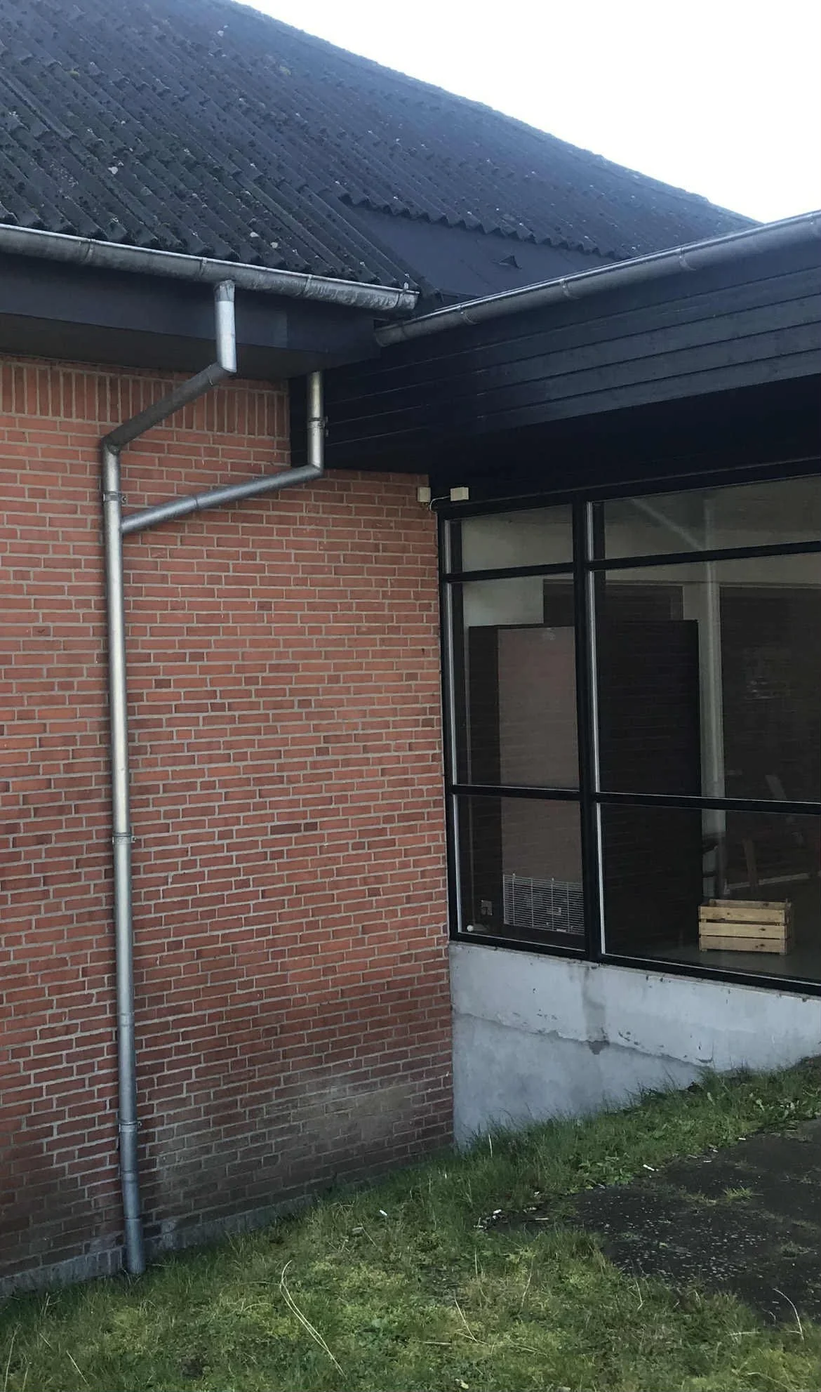 Udsigt af bygning med røde mursten, store vinduer og tag med sorte tagsten, med udendørs nedløbsrør.