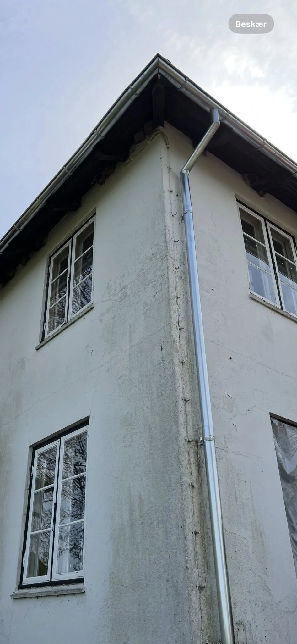 Et hjørne af et hvidt hus med fire vinduer, hvor nogle af vinduerne har skodder, og væggen viser tegn på snavs og revner. Der er en metal nedløbsrør, der løber ned ad hjørnet, og taget er synligt, med tagrender i metal på taget.