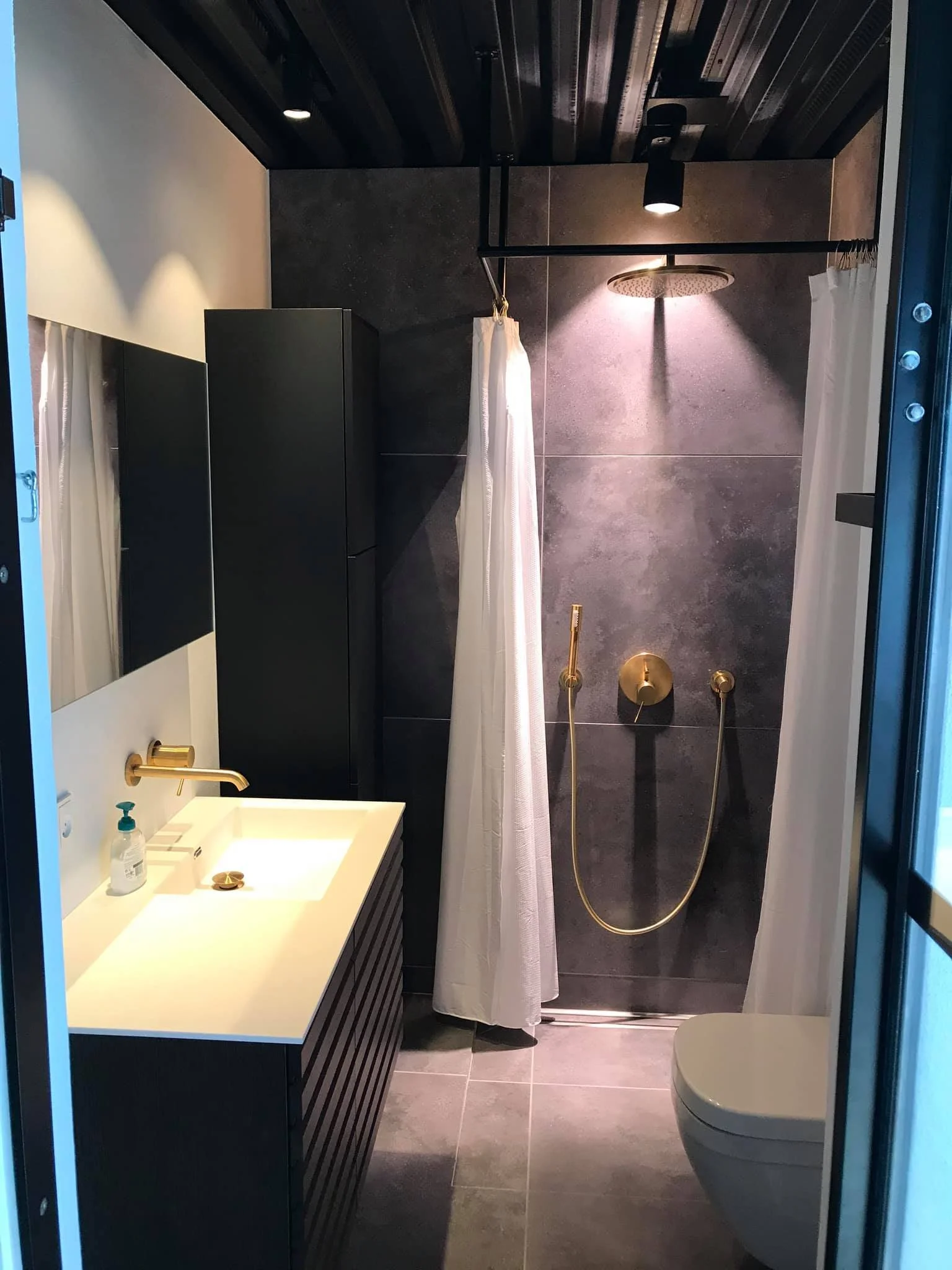 Moderne badeværelse med sort væg og hvide elementer, herunder vask med håndsæbe og spejl, bruseniche med guldkantet bruserarmatur, brusehoved og gardin, toilet, træloft og gulv, dæmpet belysning.