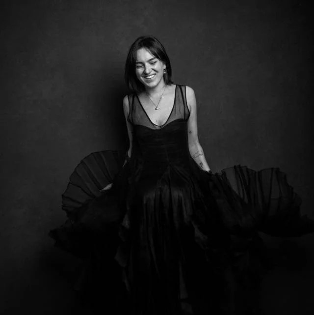 A smiling woman with shoulder-length dark hair in a black dress sitting against a dark background.