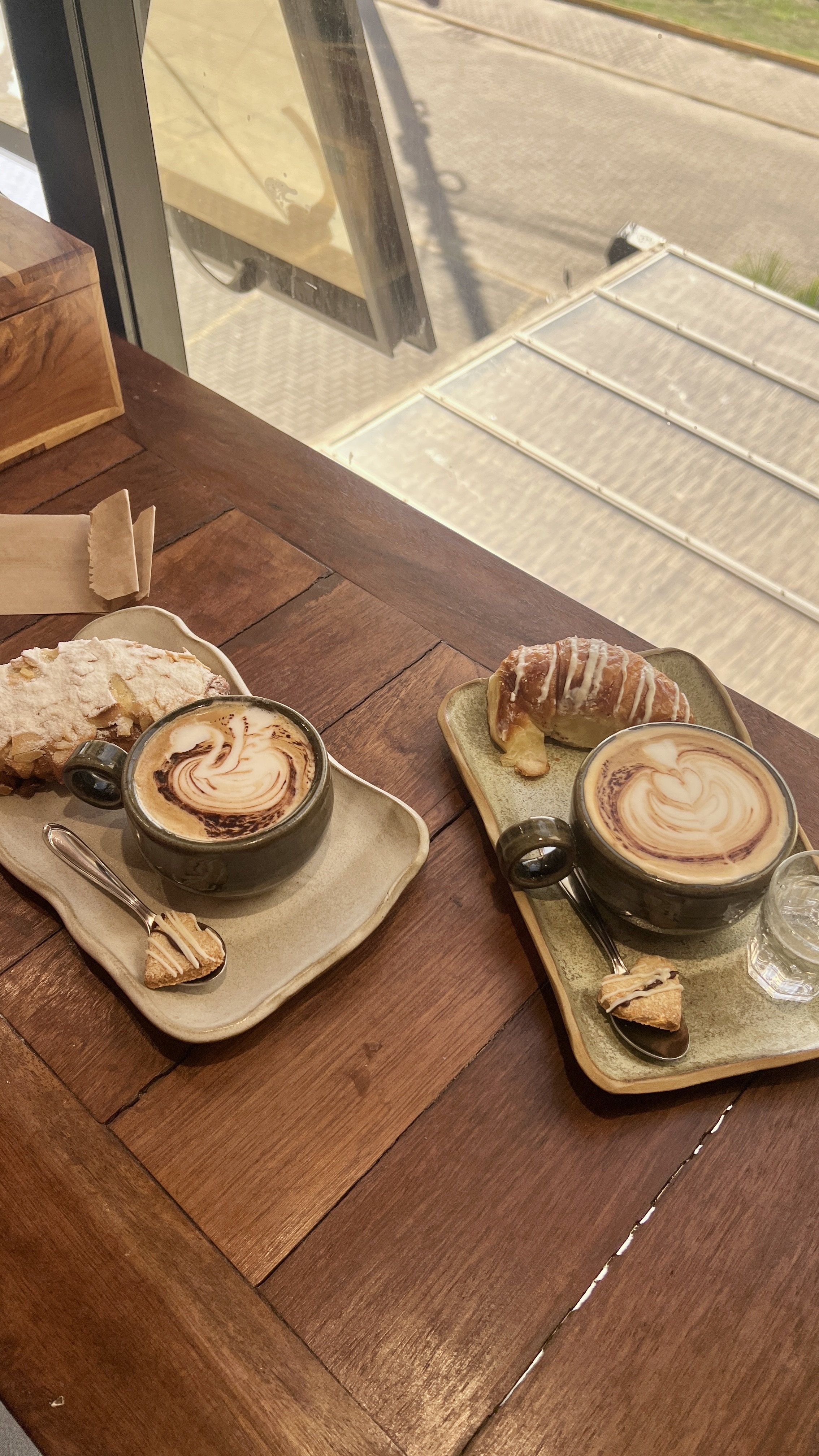 Two cups of coffee with latte art, two pastries on small plates, a glass of water, and a spoon on a wooden table near a window. A client at a menopause support consultation
