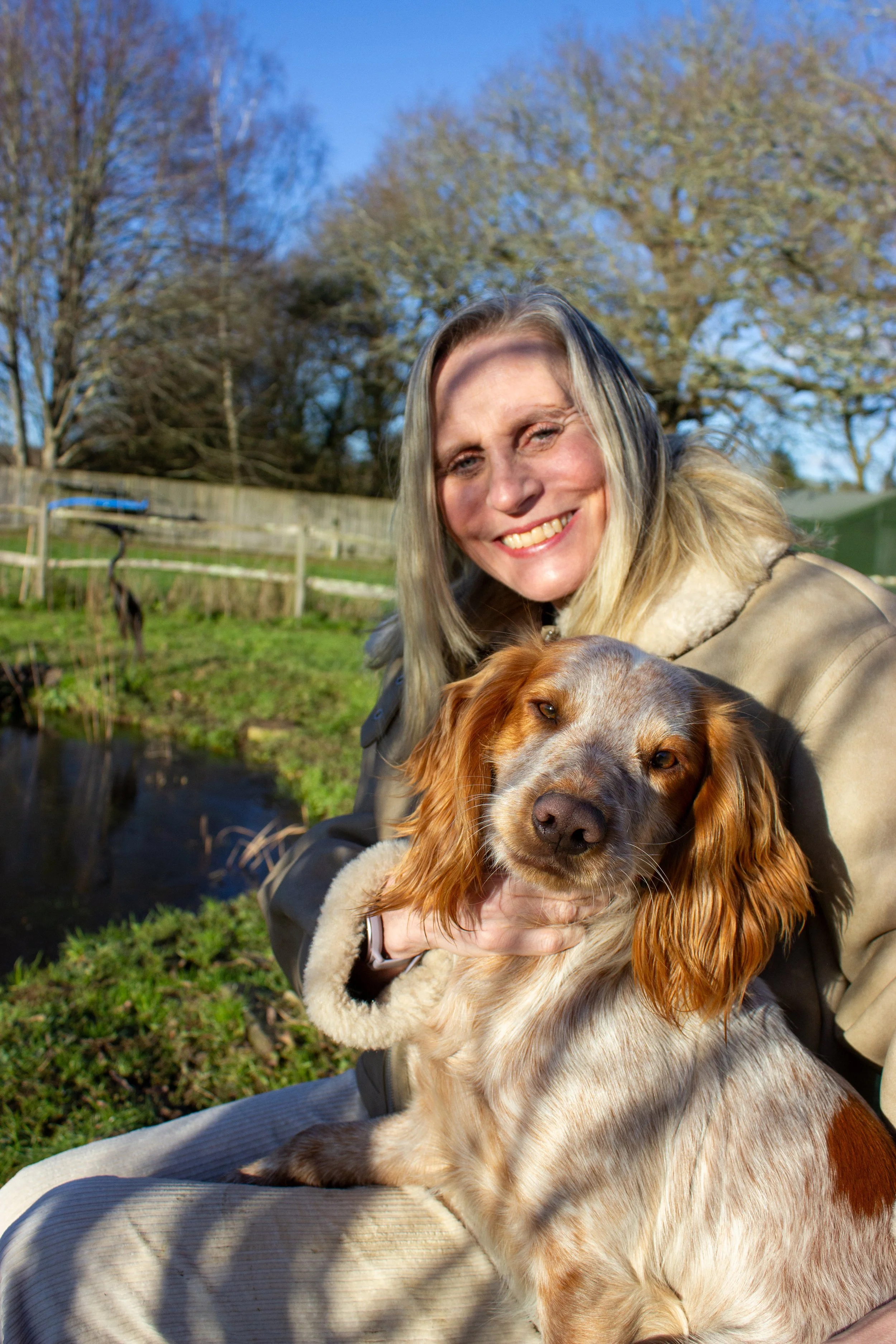 A woman, Caroline a certified menopause coach in salisbury Wiltshire, smiling while holding her dog outdoors on a sunny day, with trees and a wooden fence in the background.