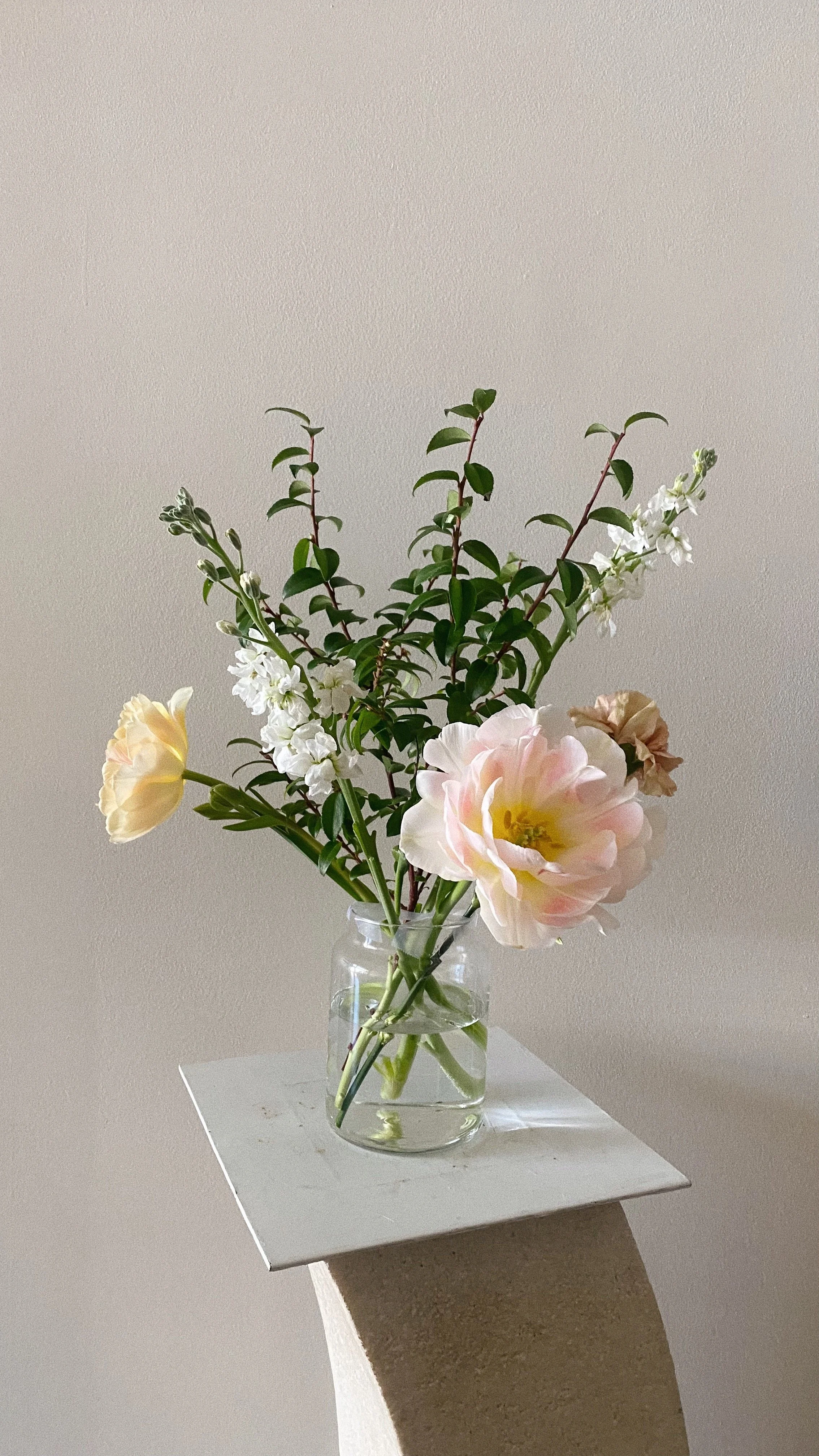 A glass vase with pink, white, and cream roses, along with green foliage, is placed on a white pedestal against a plain light-colored wall in the menopause coaching office.