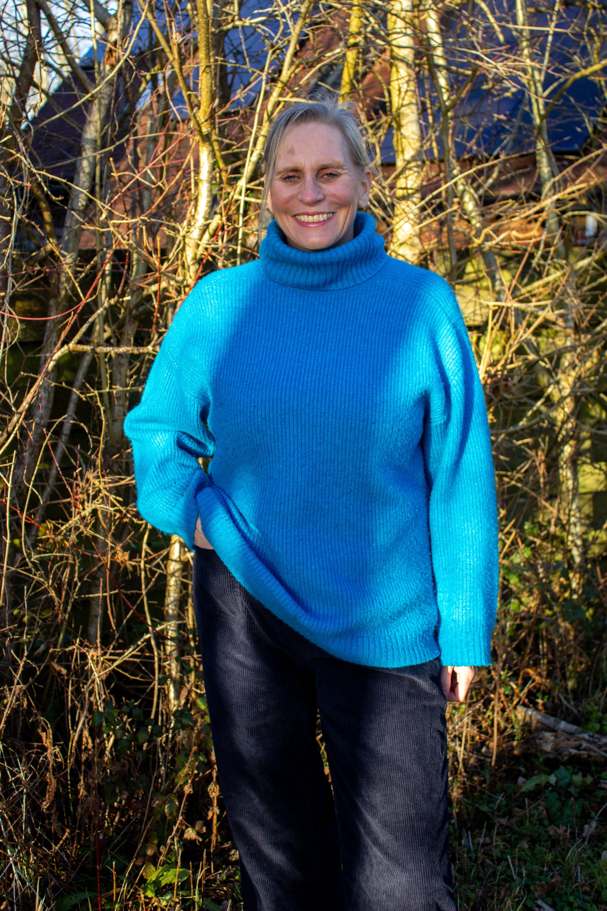 Caroline the menopause workshop mentor with light hair, smiling, wearing a bright blue turtleneck sweater and dark pants, standing outdoors in front of leafless trees and bushes during sunlight.