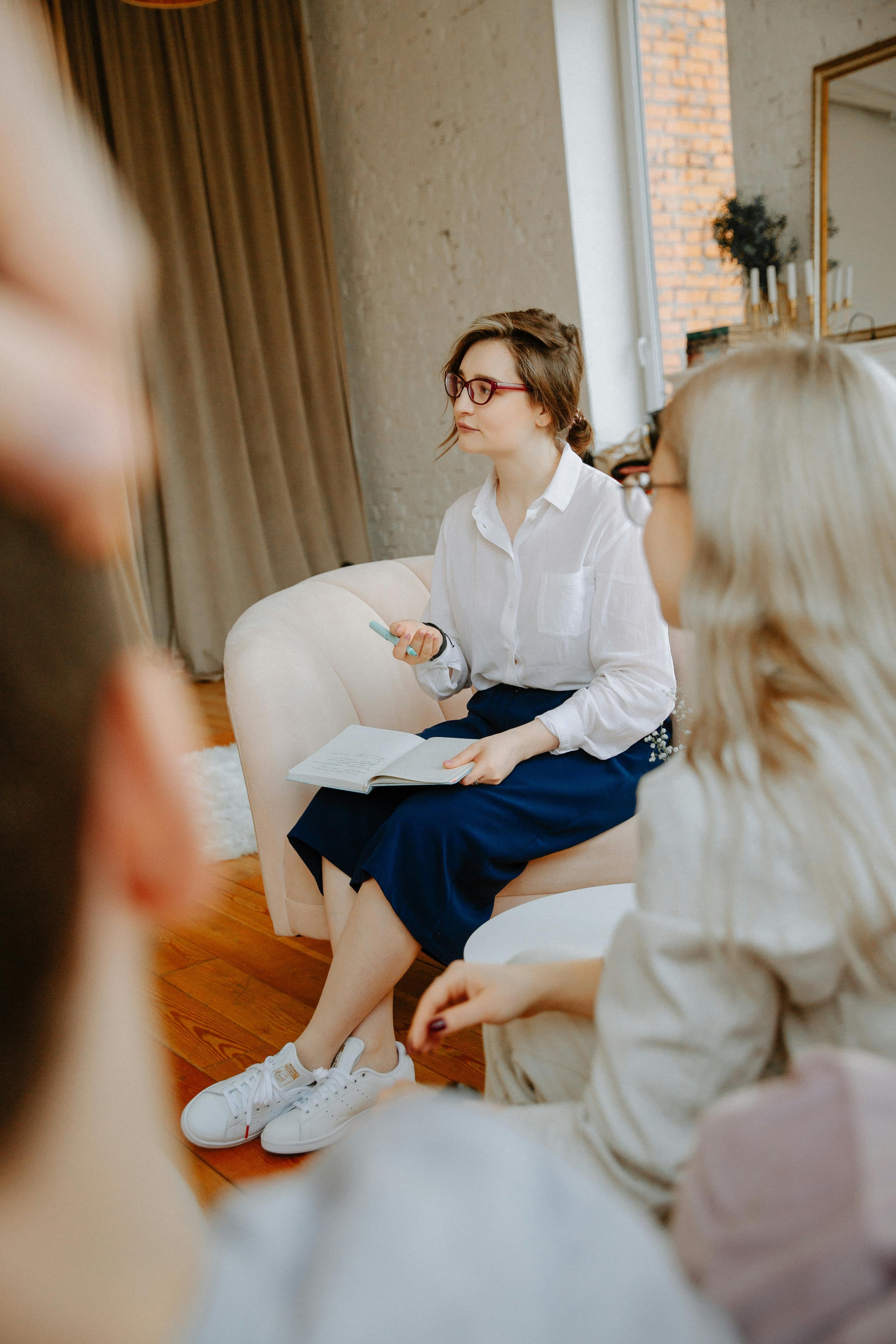 Young woman with short brown hair and glasses, sitting on a light-colored sofa, holding a notebook and a pen, engaged in conversation at a menopause awareness training day at work led by a menopause coach.
