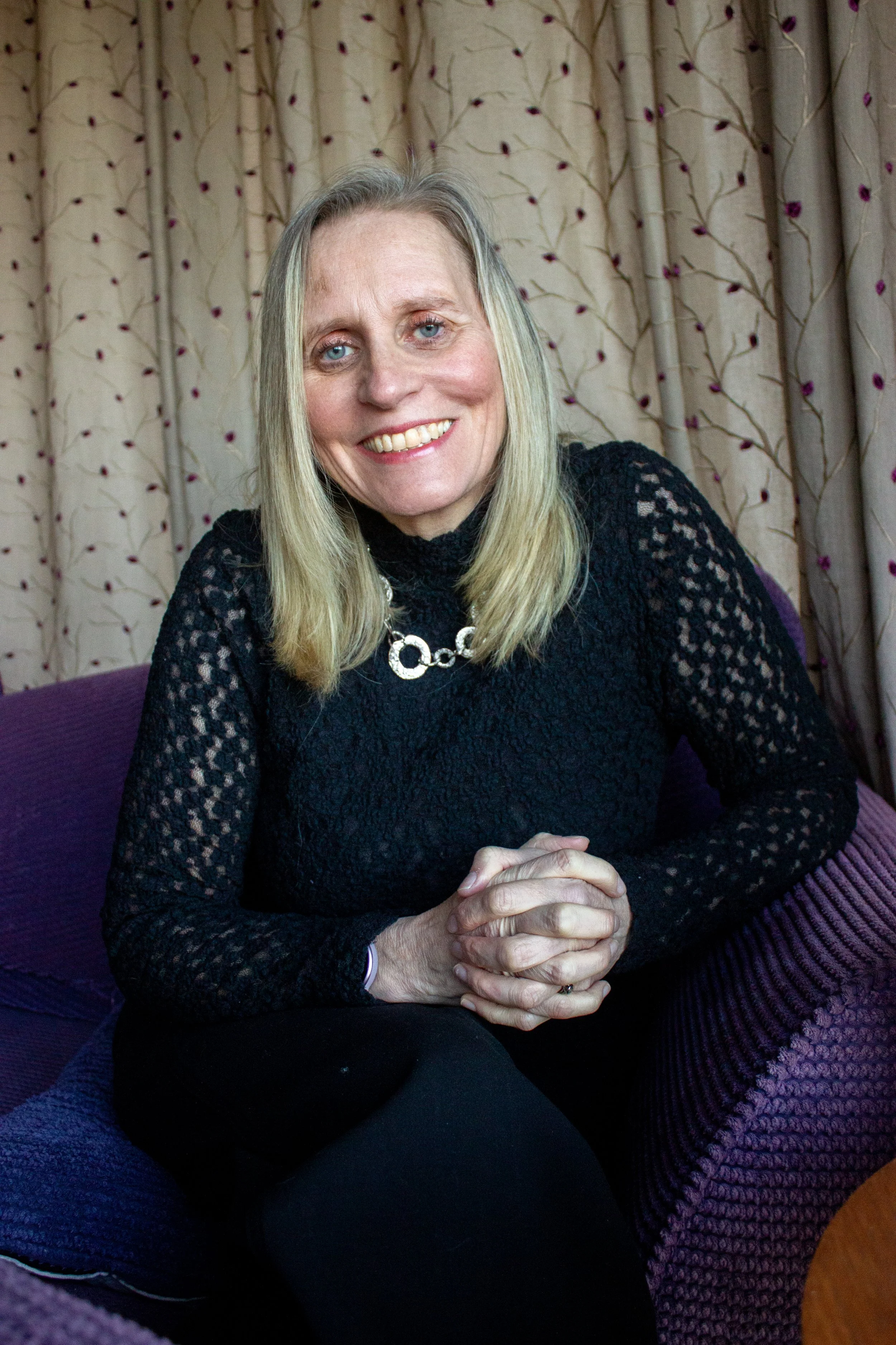 Caroline, certified menopause coach and mentor in Salisbury, smiling woman with blonde hair and blue eyes sitting on a purple armchair with patterned curtains in the background.