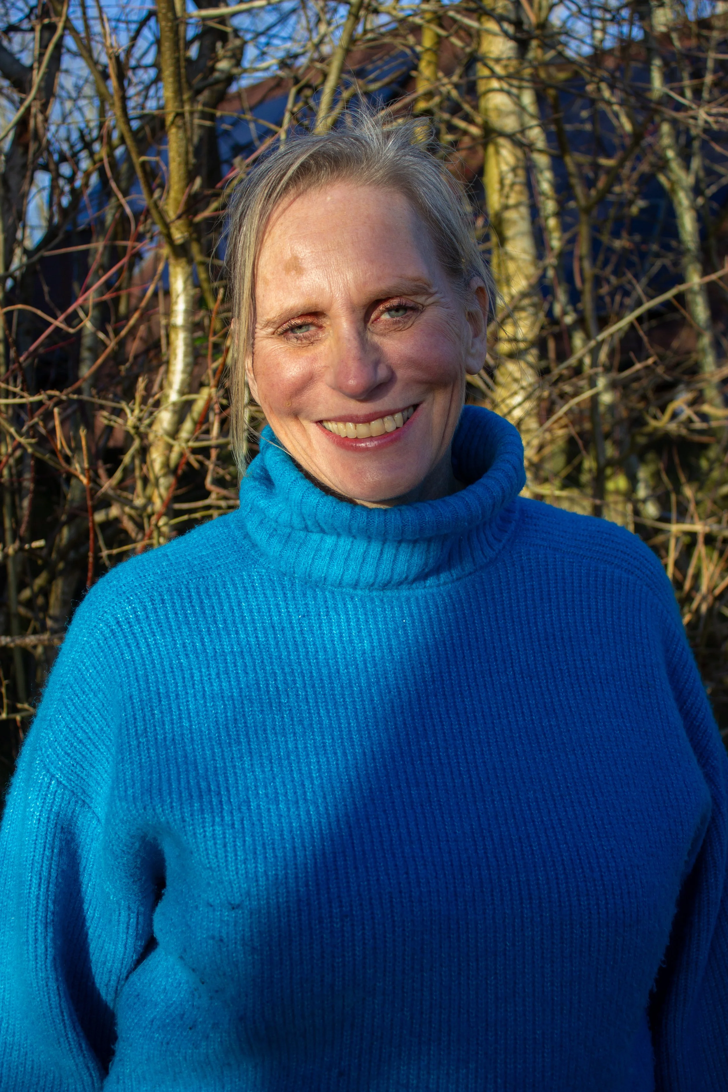 A woman in a bright blue turtleneck sweater smiling outdoors with leafless trees in the background Caroline a menopause mentor