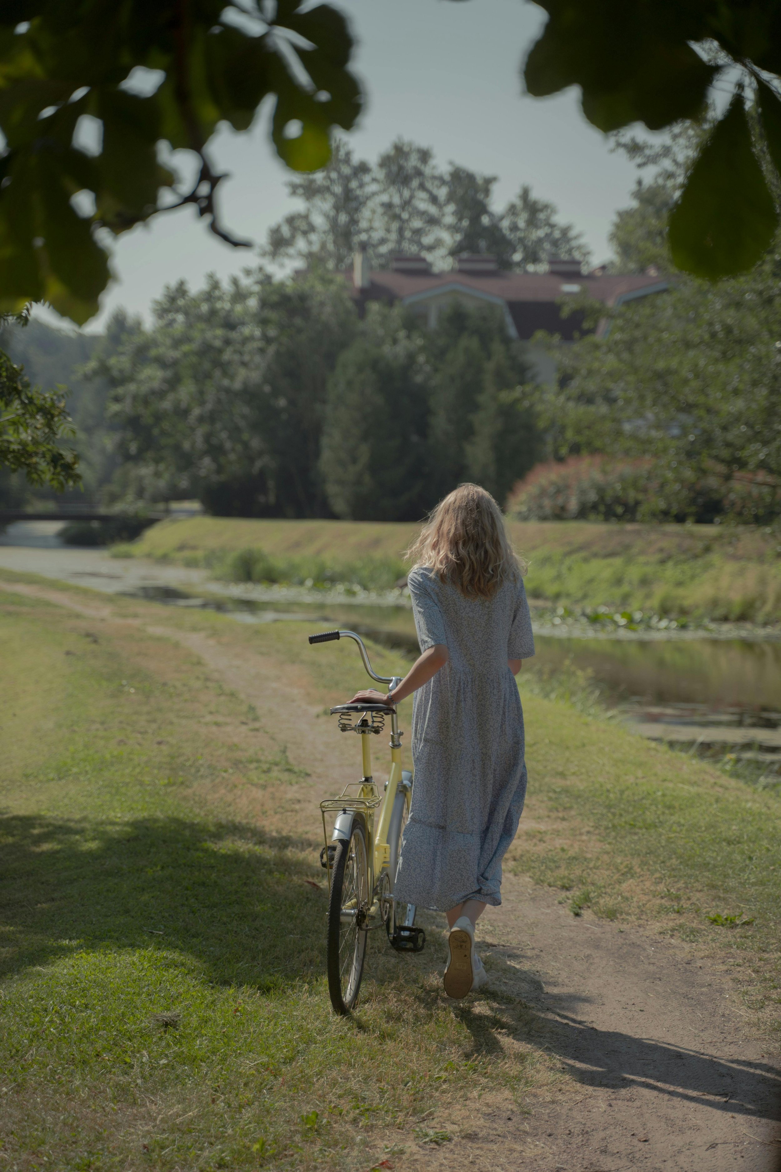 A woman at menopausal age with blonde hair wearing a long, light-colored dress pushes a yellow bicycle along a dirt path beside a river, with trees and houses in the background on a sunny day.