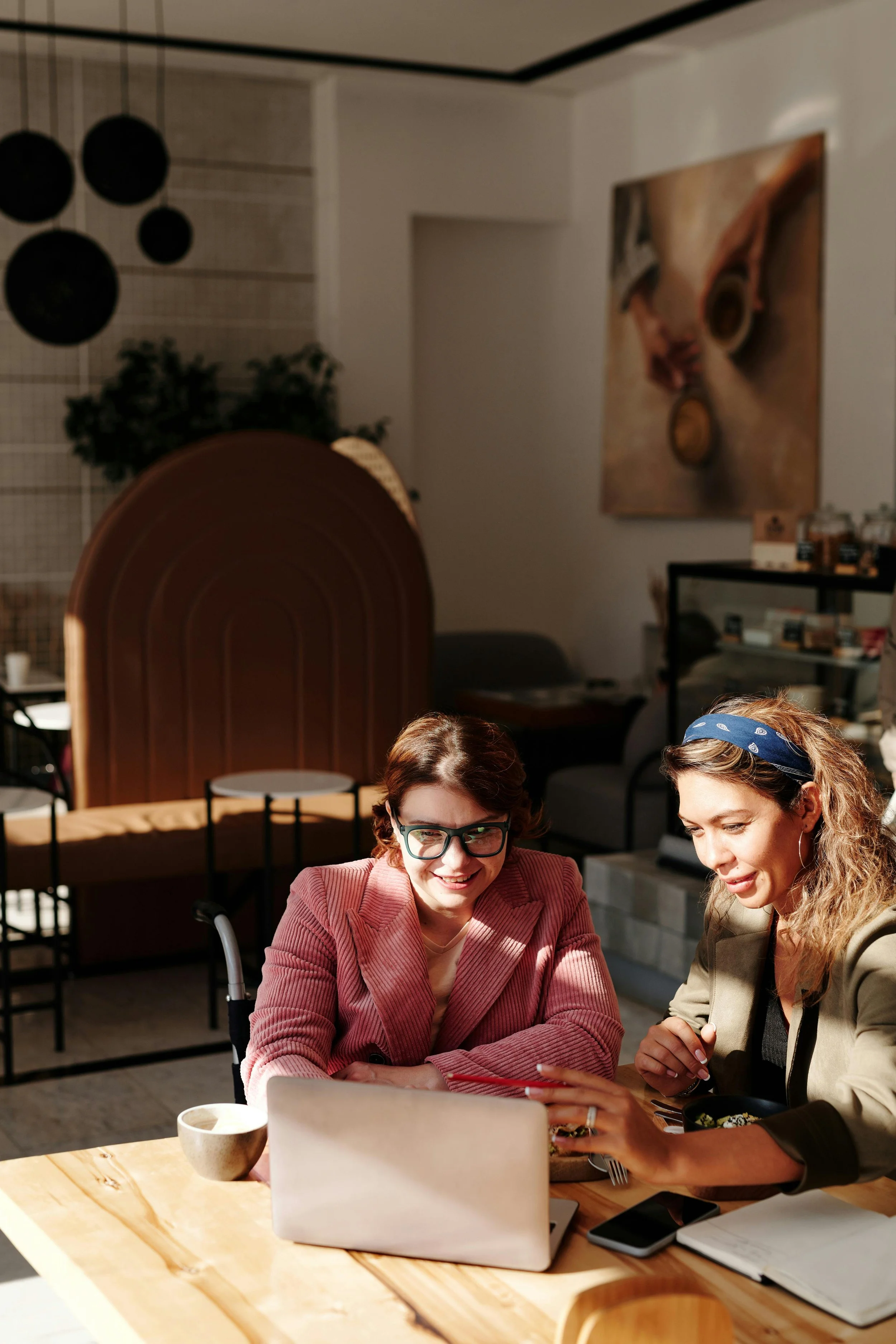 Two women sitting at a table, looking at a laptop and smiling, in a cozy cafe or workspace discussing how to reduce menopause symptoms