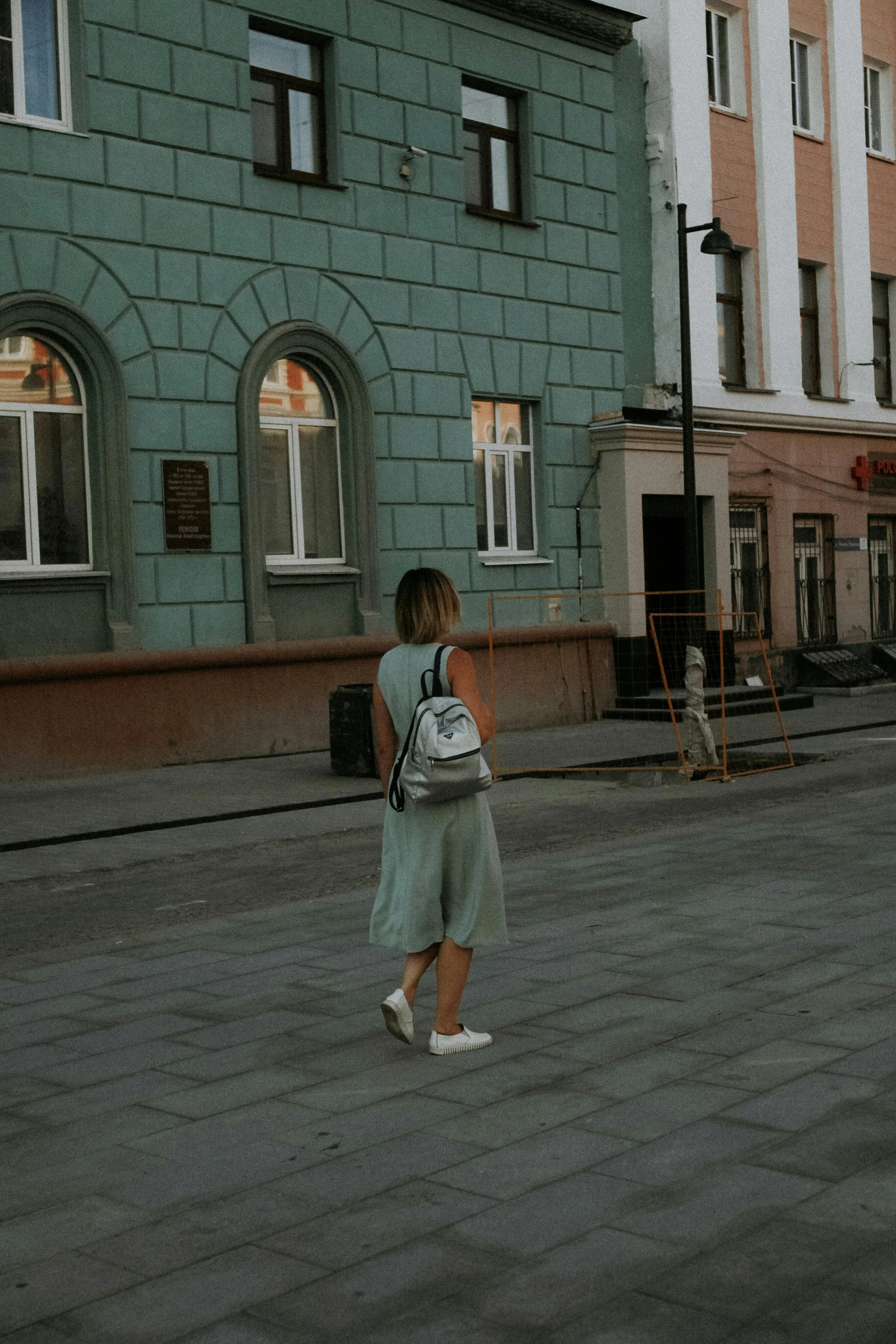 A woman struggling through the menopause with a backpack walking on a city sidewalk near pastel-colored buildings on her way to her menopause coaching appointment