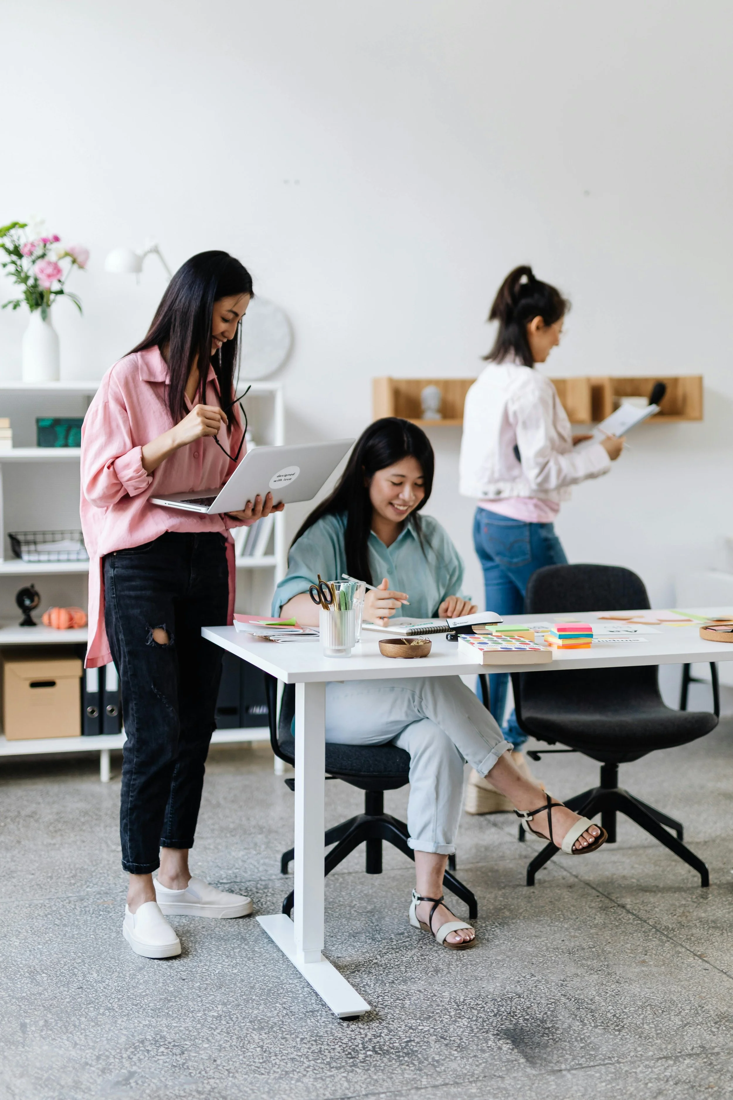Three women in an office working and chatting, one woman is sitting at a desk with stationery, and two women are standing, one with a laptop and the other with a notepad getting menopause support in the workplace salisbury