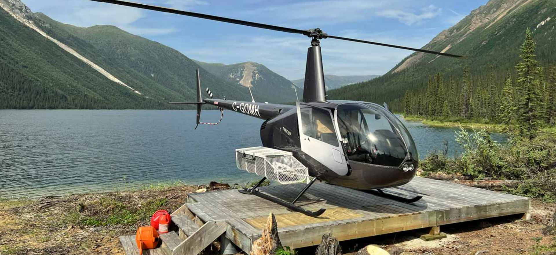 Un hélicoptère noir et blanc garé sur une plateforme en bois au bord d'un lac entouré de montagnes et de forêt, sous un ciel bleu