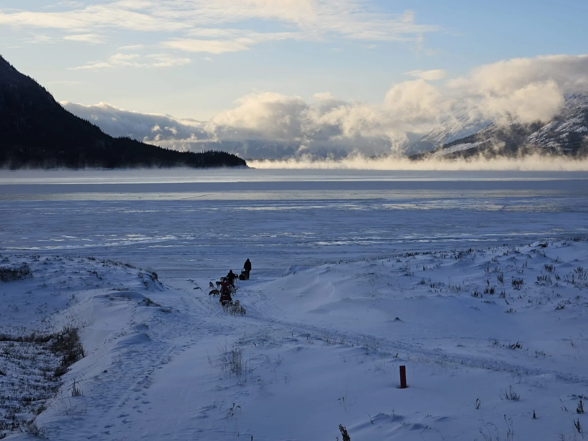 Paysage hivernal avec des montagnes enneigées, un grand lac gelé et des traces dans la neige avec des chiens tirant un traîneau, au centre, avec un ciel partiellement nuageux juste sur le haut des montagnes du Yukon au Canada.