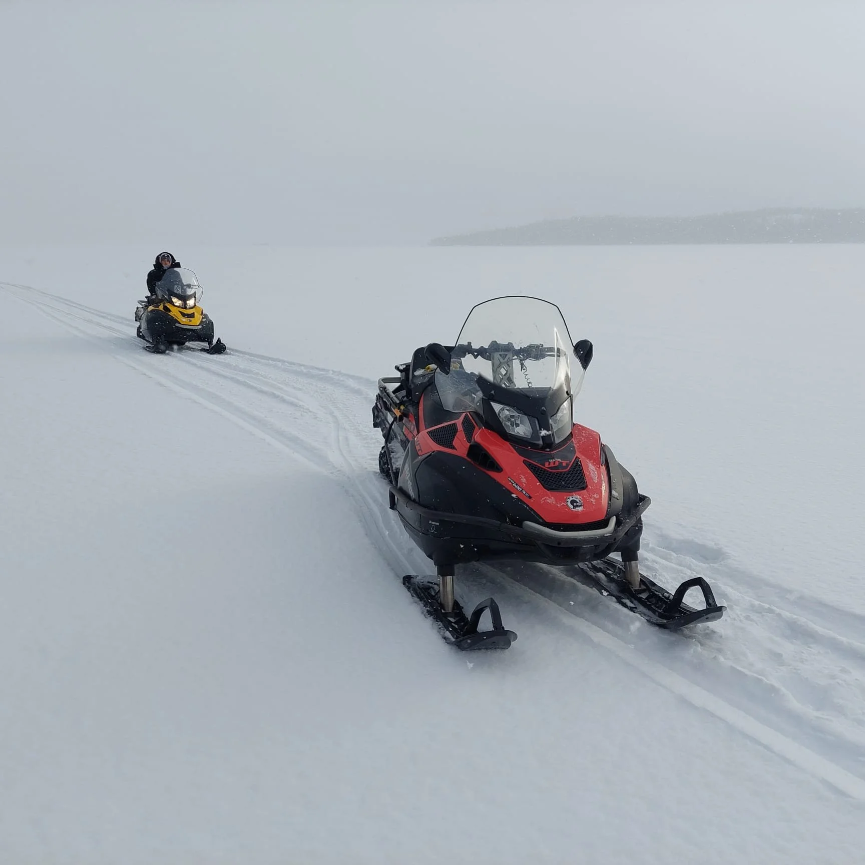 Gros plan d'un motoneige rouge sur une surface enneigée, avec une autre motoneige en arrière-plan dans un paysage neigeux brumeux tôt le matin d'une excursion dans le Yukon.