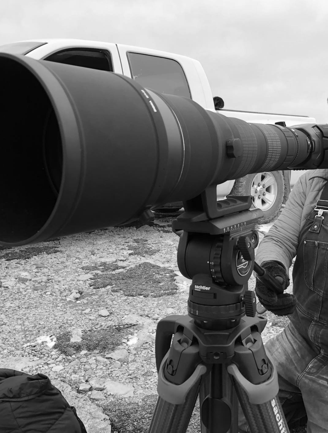 Photo en noir et blanc d’un télescope avec un support sur un trépied, un homme en gants et veste en Wikipedia, et une voiture blanche en arrière-plan, dans un paysage rocheux.