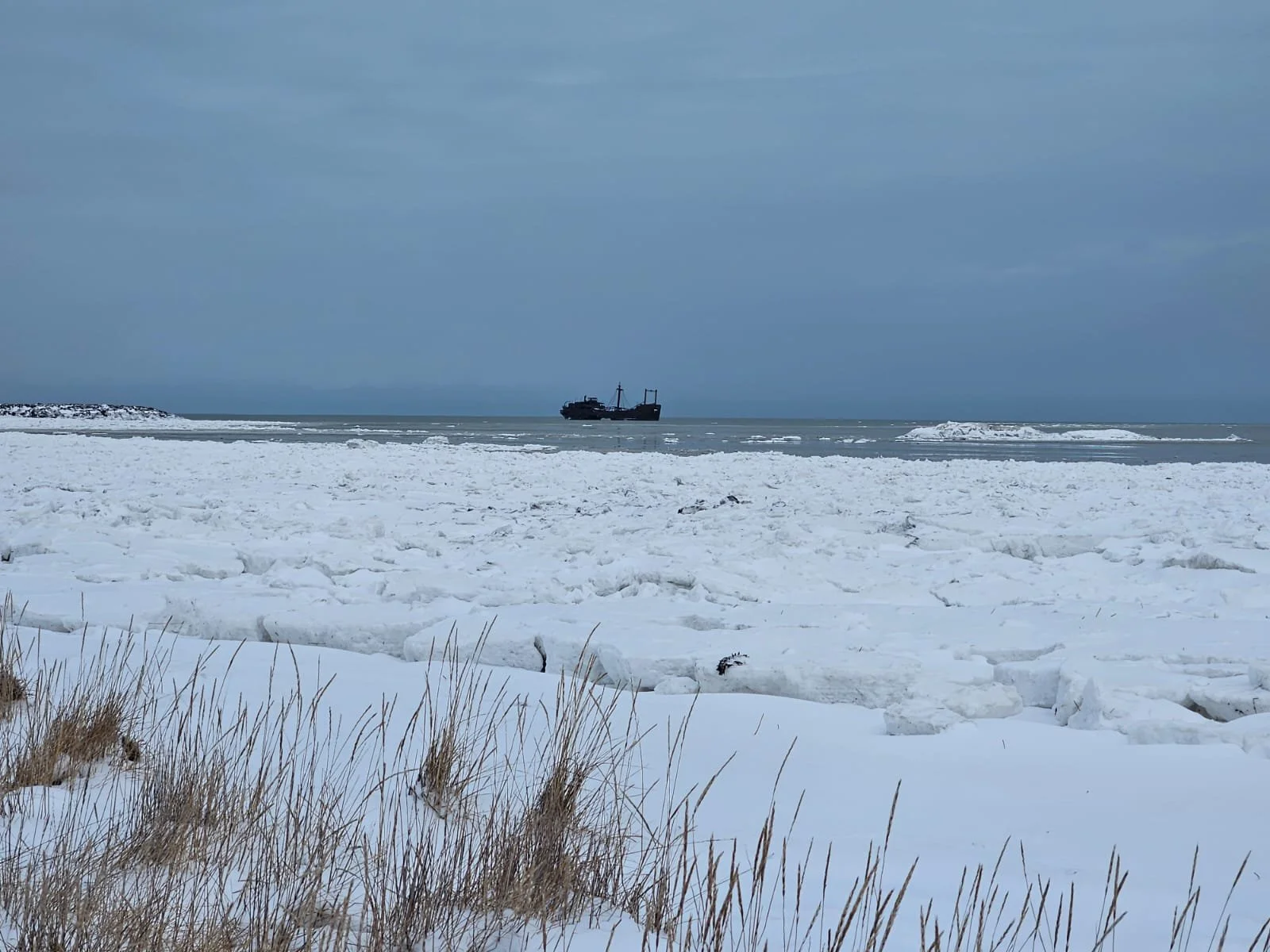 Scène glacée avec une plage recouverte de glace et de neige, un bateau rouillé en arrière-plan de la photo (le célèbre bateau de Churchill) en mer arctique, avec un ciel gris.
