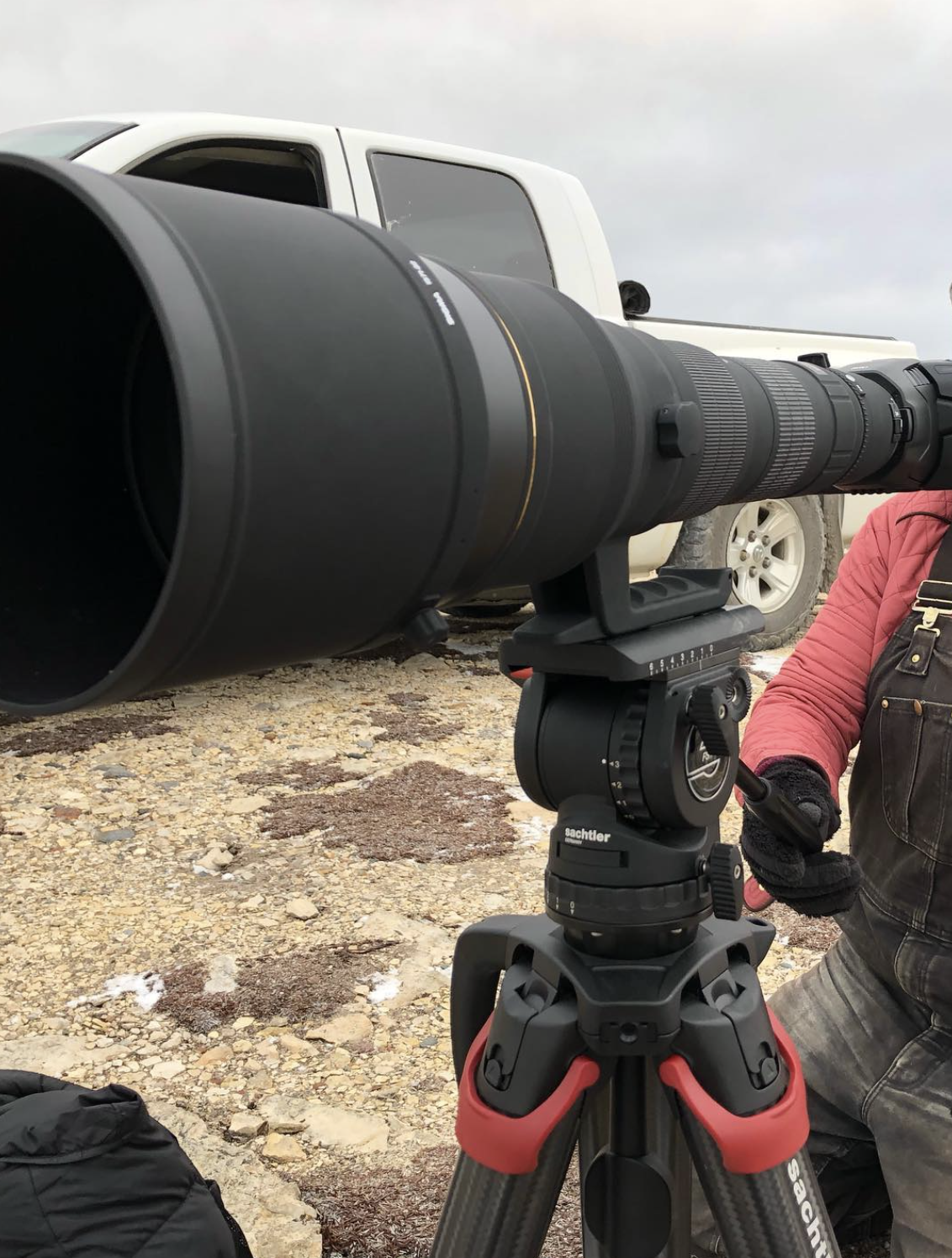 Un objectif photo professionnel monté sur un appareil avec trépied, avec une personne en vêtement chaud ajustant ou utilisant l'équipement, se trouvant à l'extérieur sur un sol rocheux avec un véhicule de type pick-up en arrière-plan.