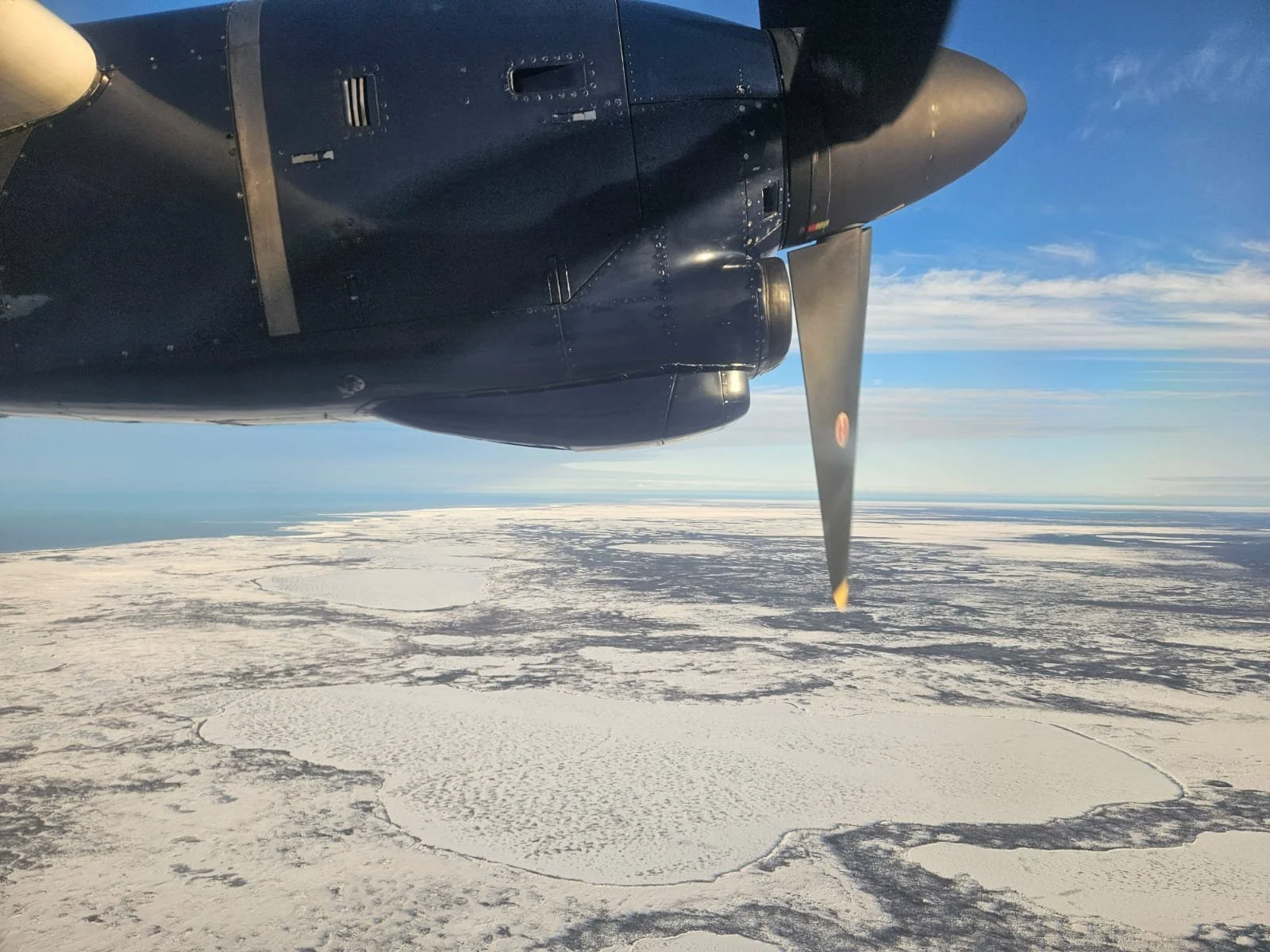 Vue depuis un avion montrant la moitié d'une hélice et un paysage glacé sous un ciel bleu avec quelques nuages.