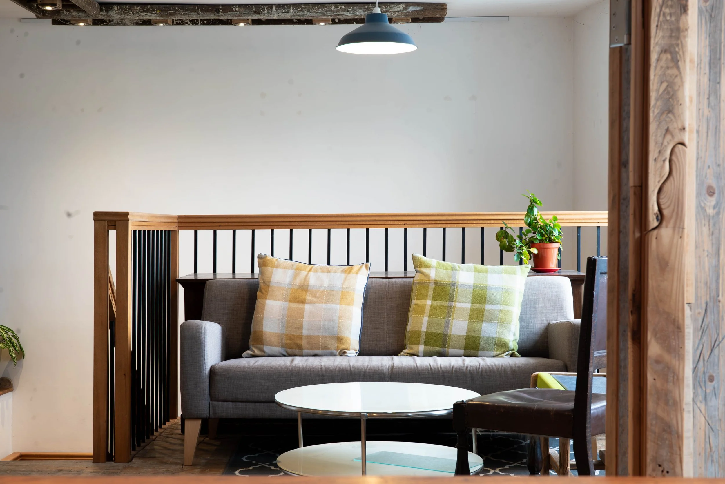 Living room with a light gray sofa adorned with two checkered pillows, a small white round coffee table in front, a wooden railing behind with black vertical balusters, a potted plant on a side table, and a dark wood chair on the right, with a ceiling light hanging from an exposed wooden beam.