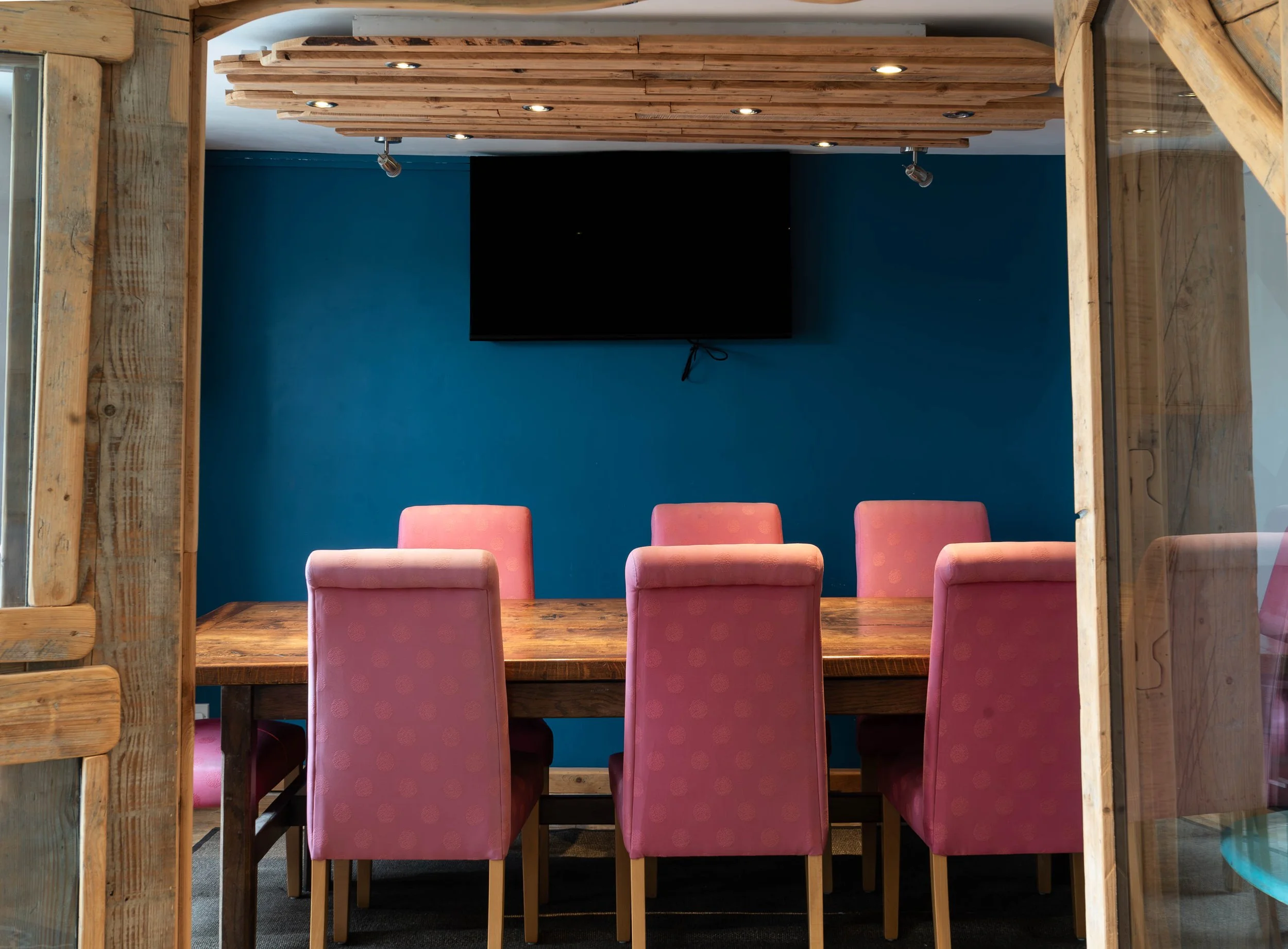 A conference room viewed through a window with wooden framing. The room has a large wooden table surrounded by six pink upholstered chairs. The wall behind the table is painted blue, and a flat-screen TV is mounted on it. Recessed ceiling lights are also visible.