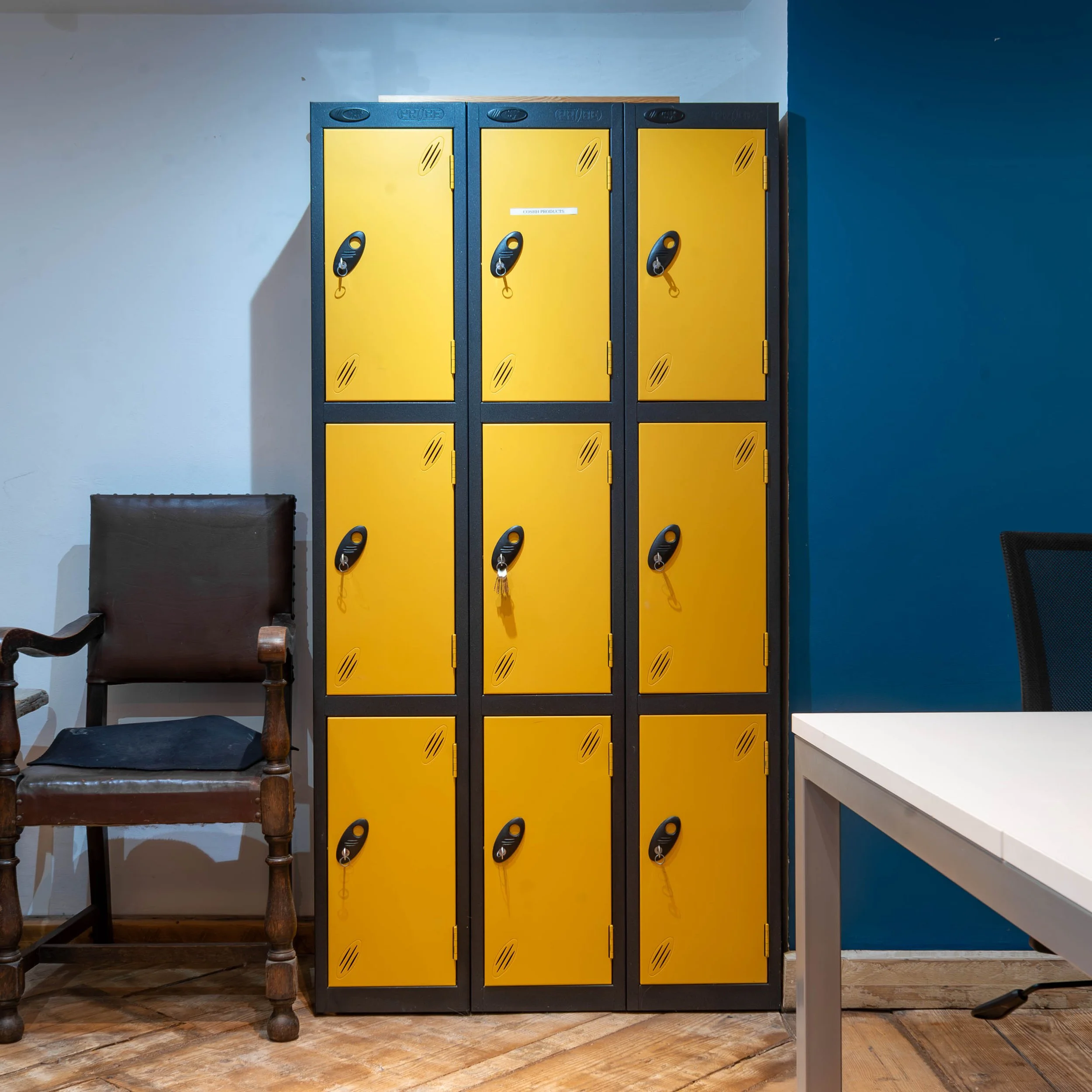 A set of nine yellow lockers arranged in three columns and three rows, with black doors and padlocks, next to a brown upholstered chair and a white table in a room with blue and white walls.