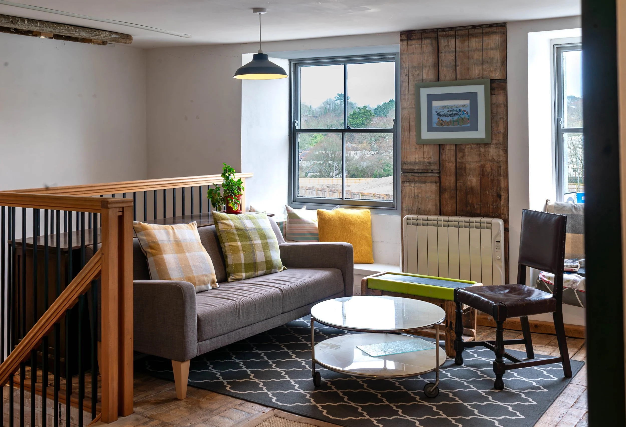 A cozy living room with a gray sofa decorated with plaid and solid pillows, a round glass coffee table on a black and white patterned rug, a wooden and iron railing, two large windows, a wooden accent wall with framed art, a radiator, a black leather chair, and various decorative plants.